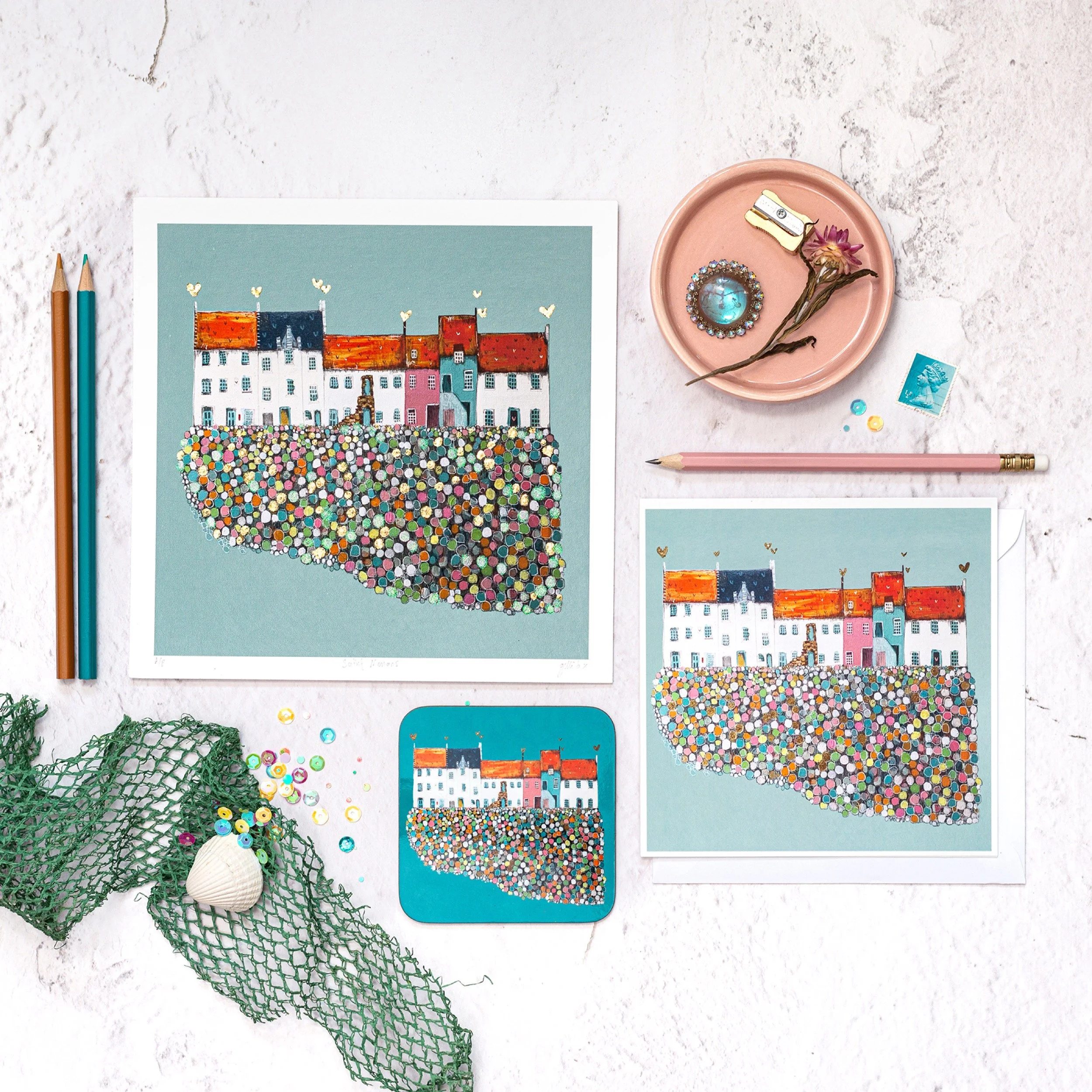 Art print of coloruful houses on the coast, with craft supplies like pencils, a pink plate, vintage brooch, and decorative items on a white surface.
