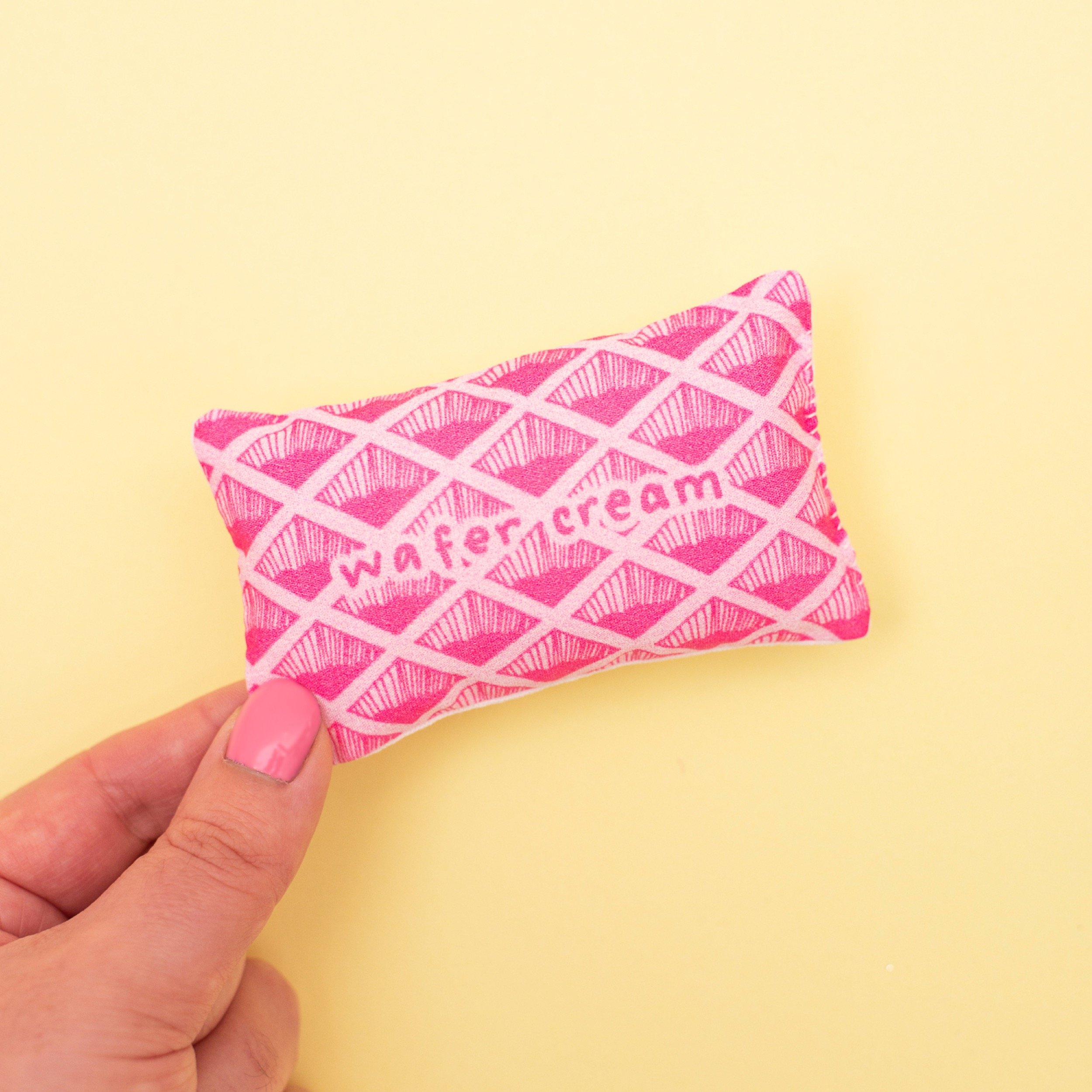 Pink 'Wafer Cream' fabric biscuit on a yellow background, held by a person's hand with pink nail polish.