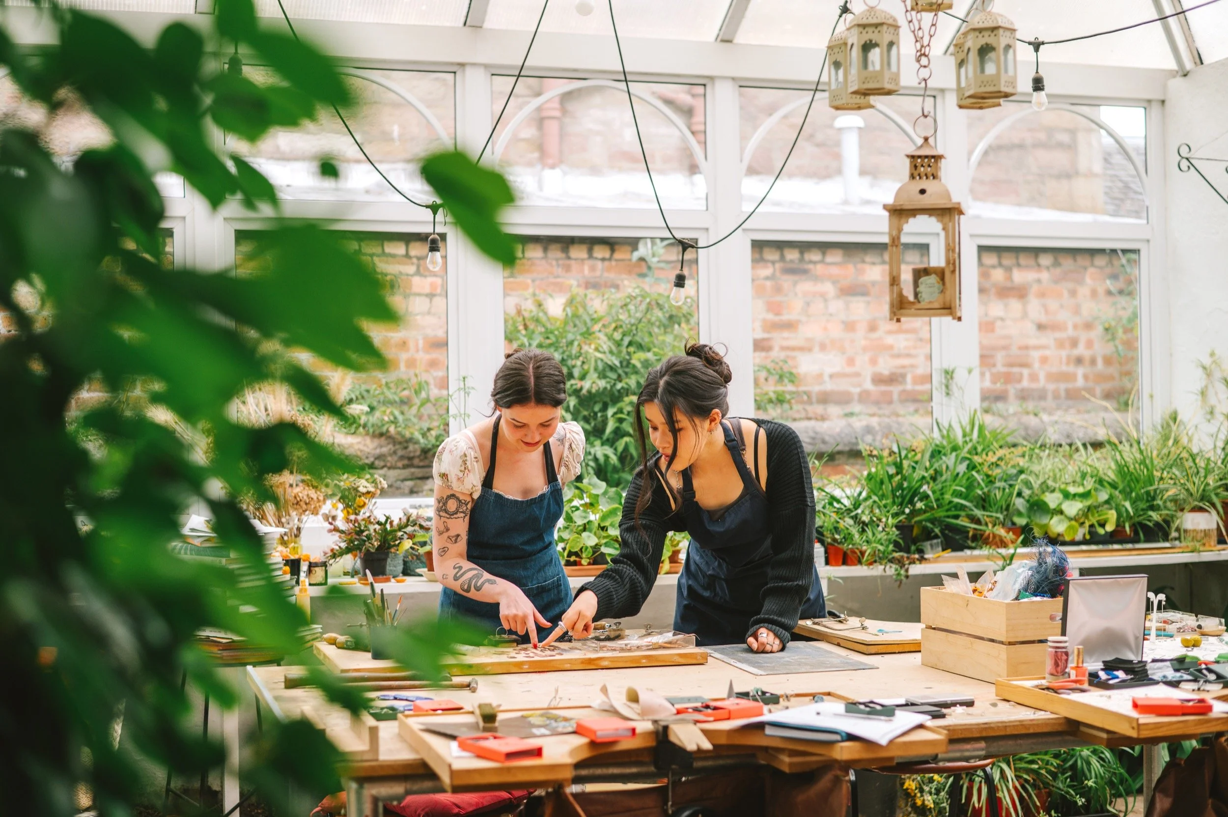 Two women working together at a jewellery bench inside an orangery, surrounded by plants, with various tools and supplies on the table.