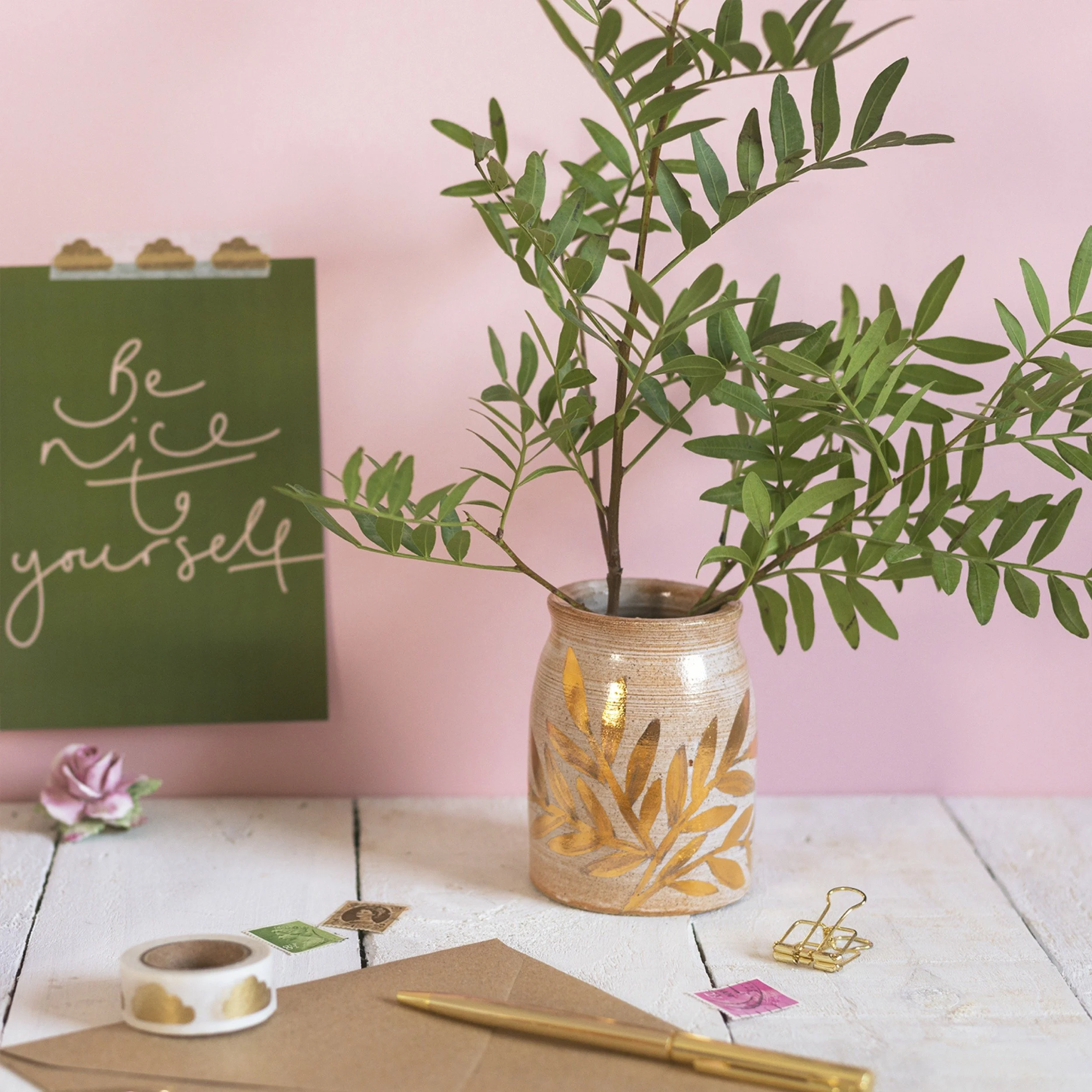 A potted plant with green leaves in a decorative ceramic vase on a white wooden surface, with a green wall and pink sign in the background that says 'Be nice to yourself,' pink flower and gold paper clips on the desk.