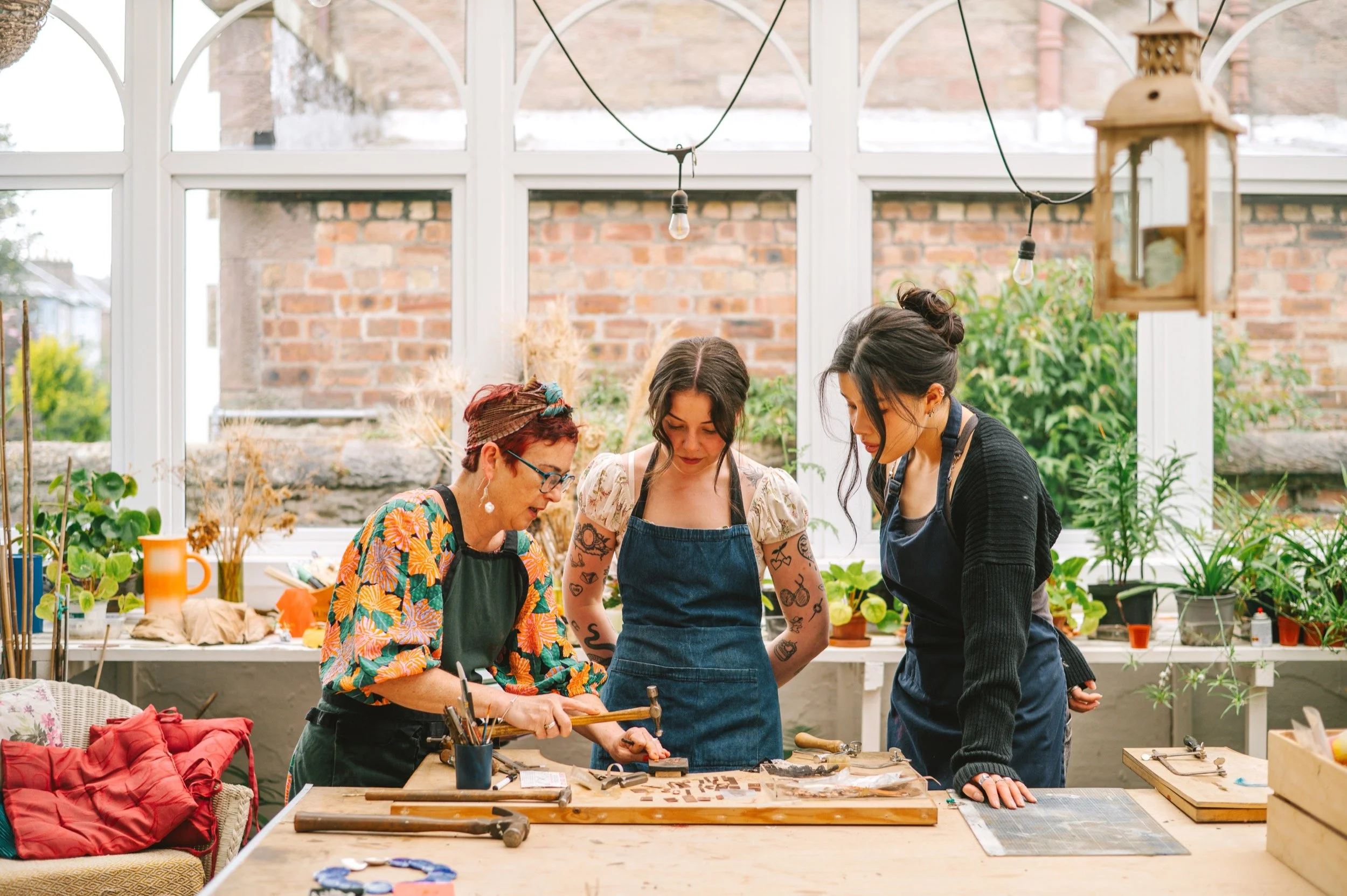 Three women working together at a jewellery bench in a bright room with large windows, plants, and brick walls.