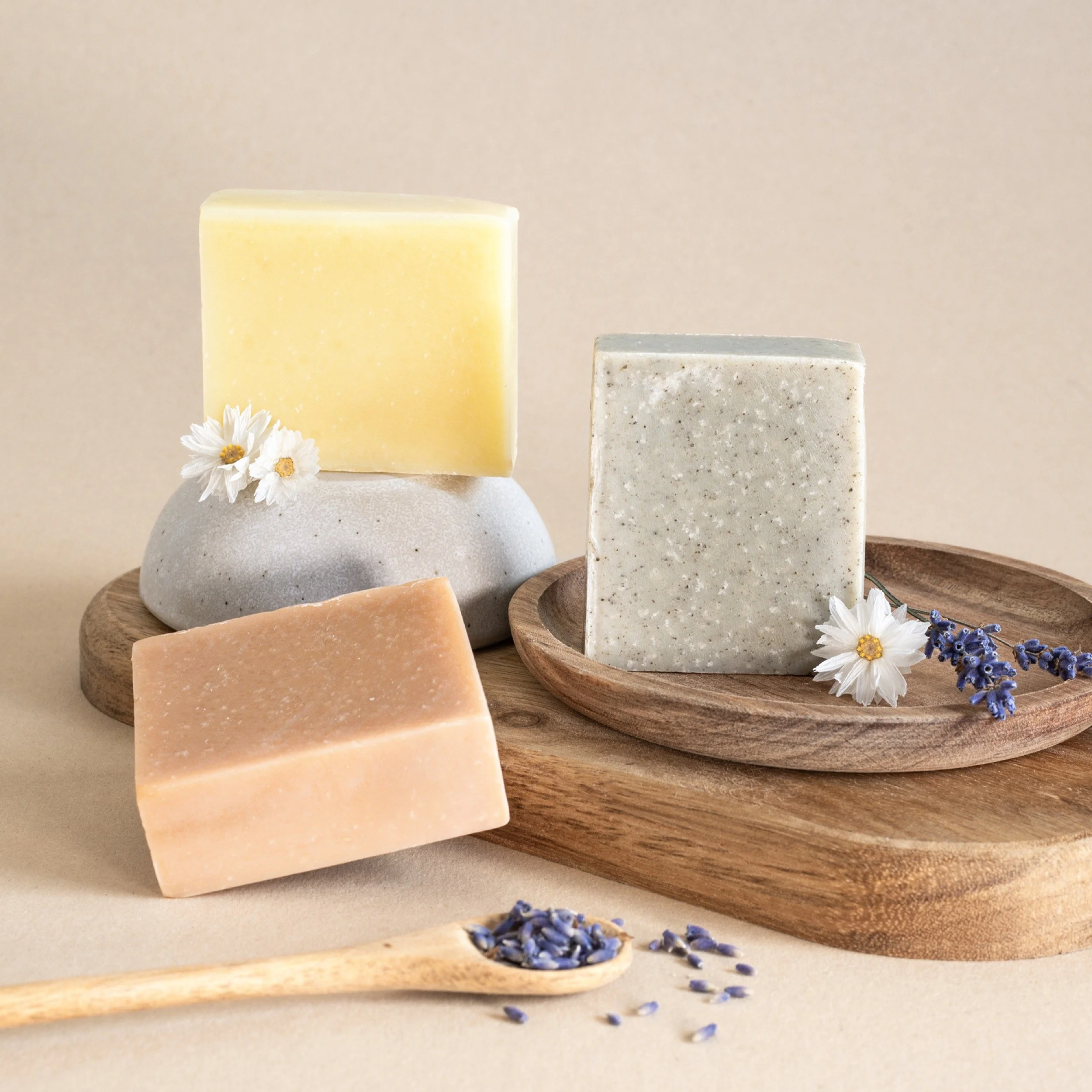 Three bars of soap in different colors and textures, with small flowers and lavender sprigs on and around wooden trays and a ceramic stand.