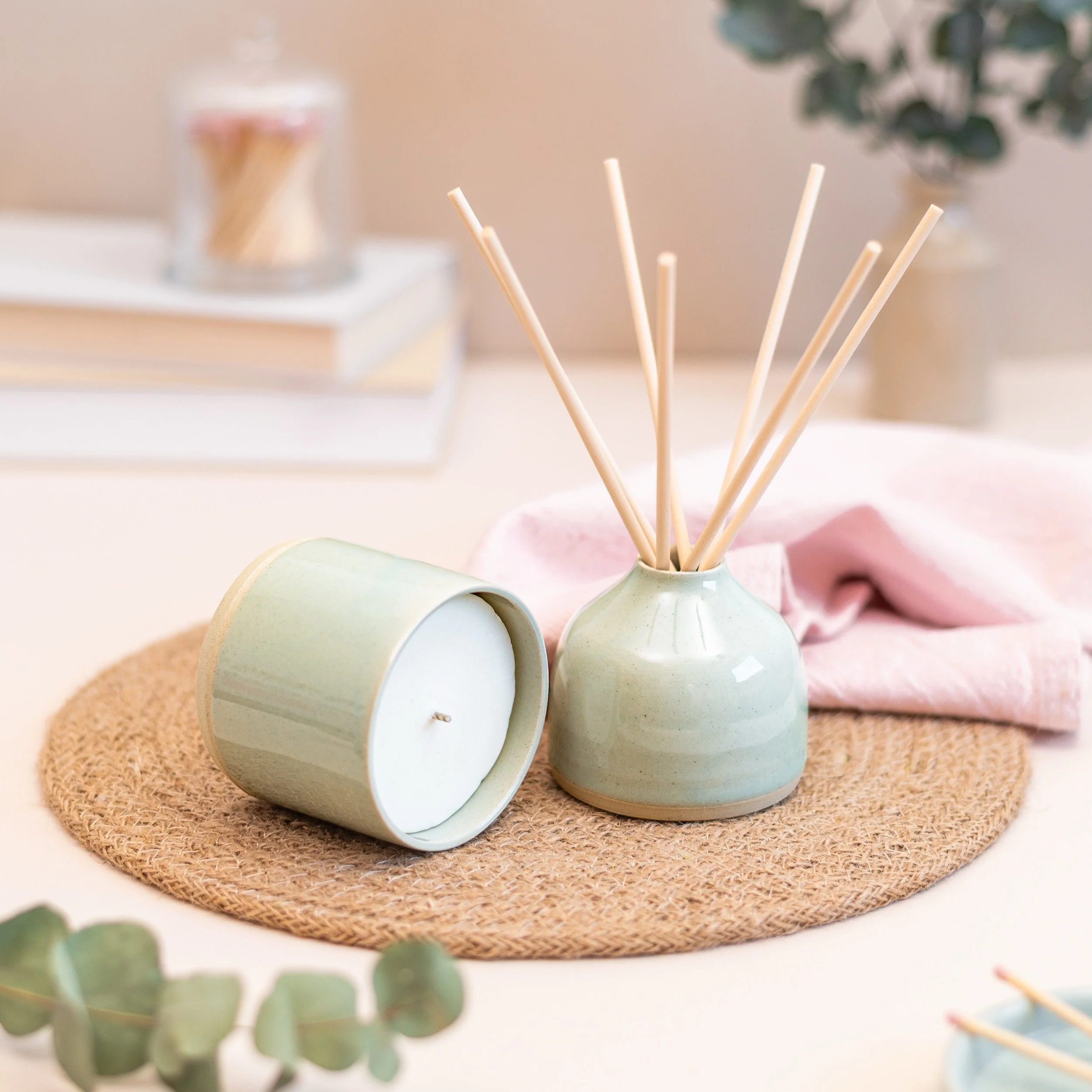 A round woven placemat with a sage green candle, a matching vase with reed diffuser sticks, and eucalyptus leaves in the foreground, set on a white surface with a blurred background.