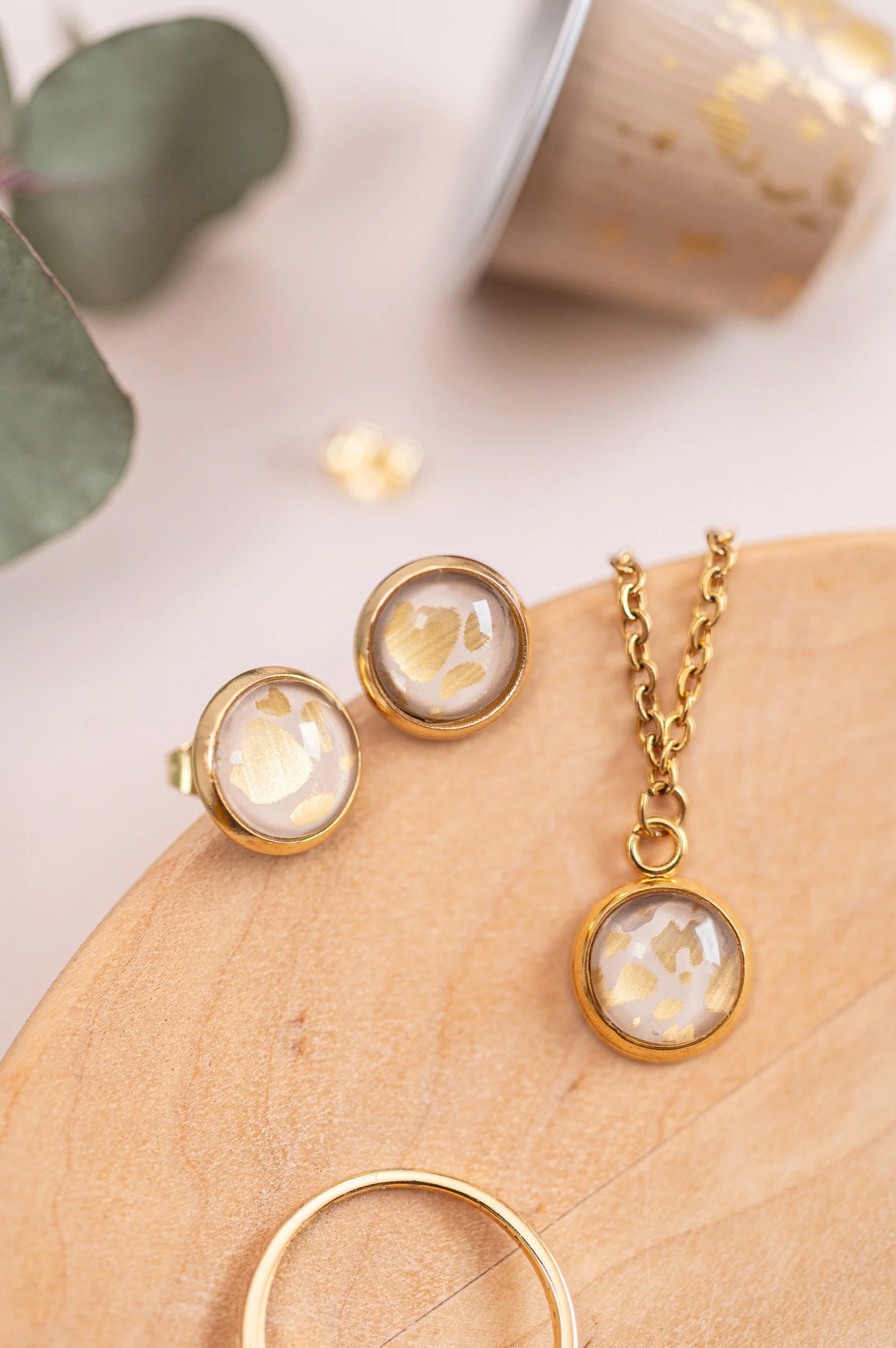 Gold jewelry set including earrings, a necklace, and a ring with circular, patterned stones on a wooden surface. In the background, there are coffee pods.