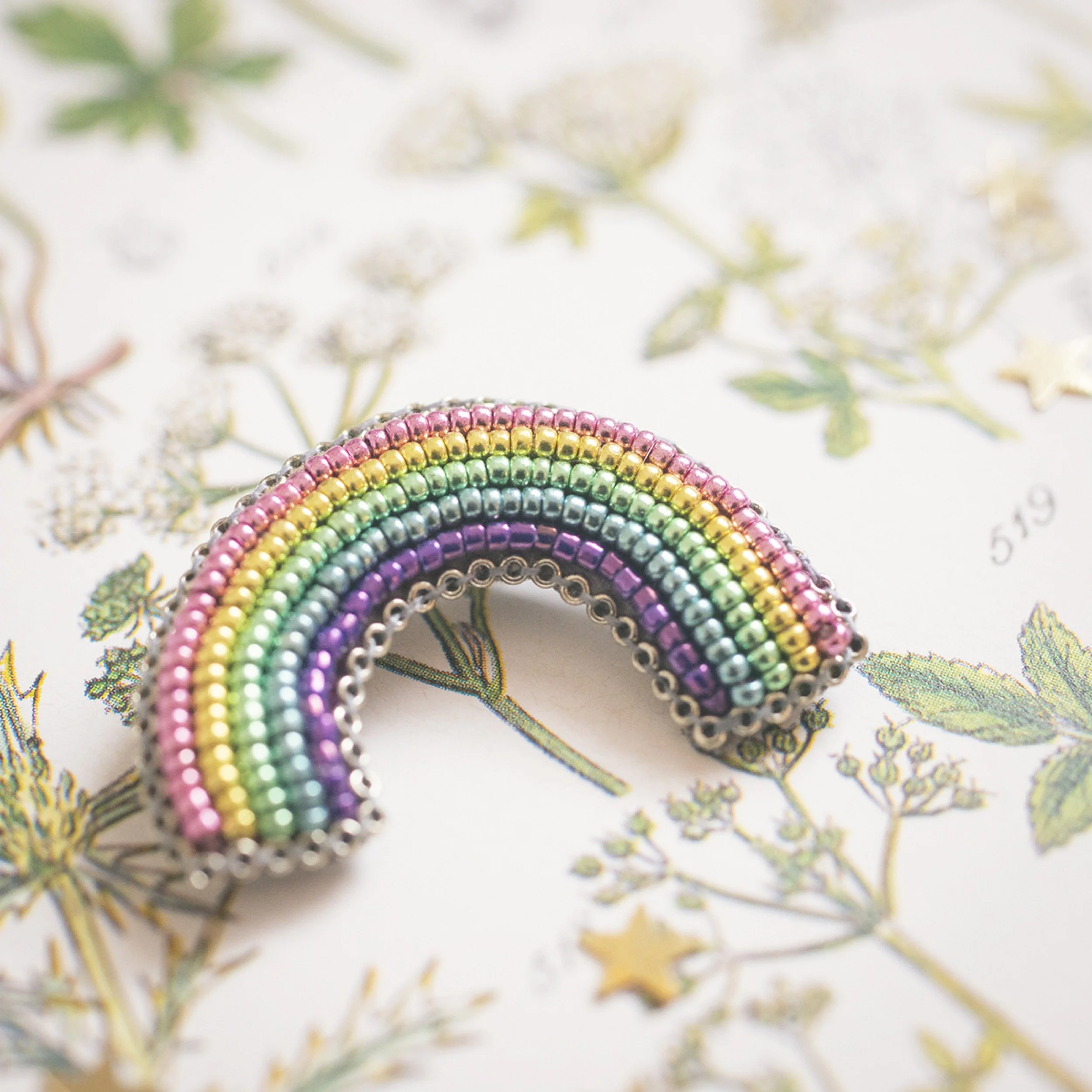 A colourful rainbow-shaped brooch made of small beads in pink, yellow, green, purple, and blue, resting on a sheet of botanical illustrations.
