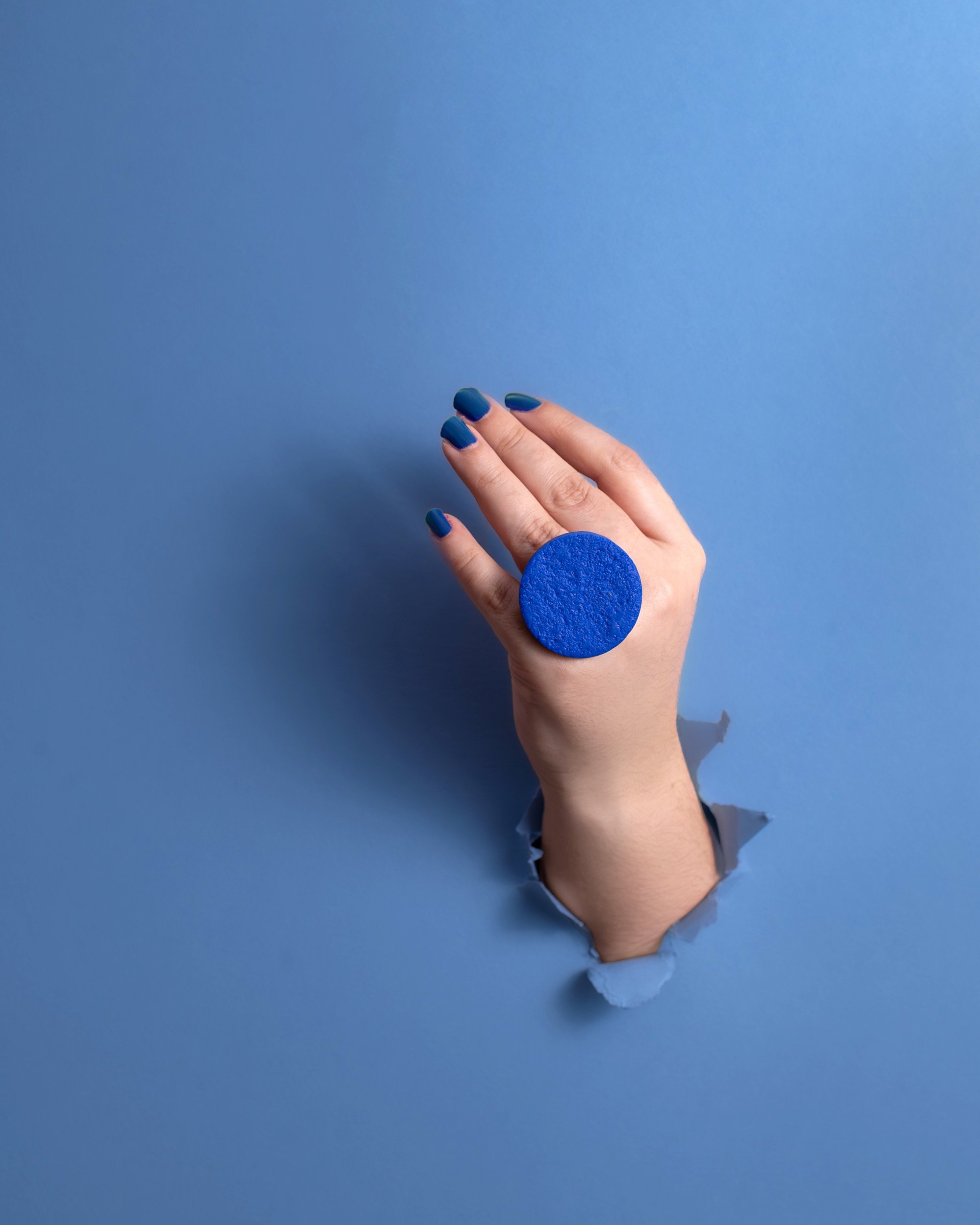 A hand with blue painted nails emerging through a tear in a blue paper background, wearing a large blue ring.