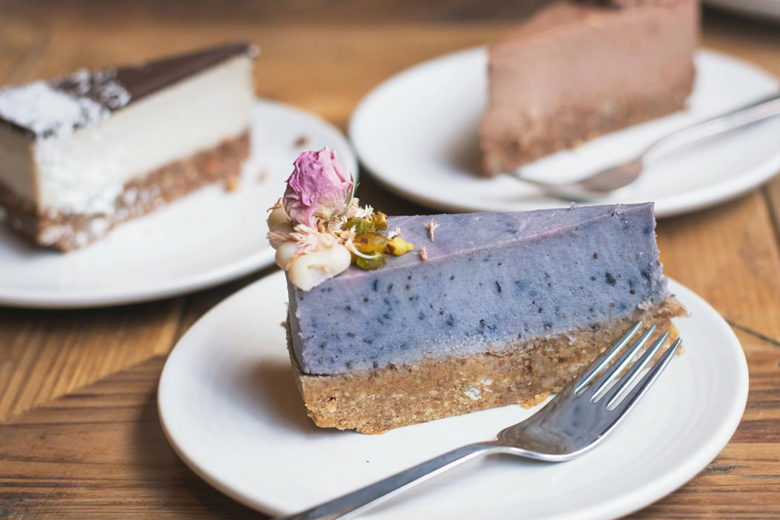 A slice of vegan cheesecake garnished with pink and white frosting flowers on a white plate, with two other cake slices on separate plates in the background.