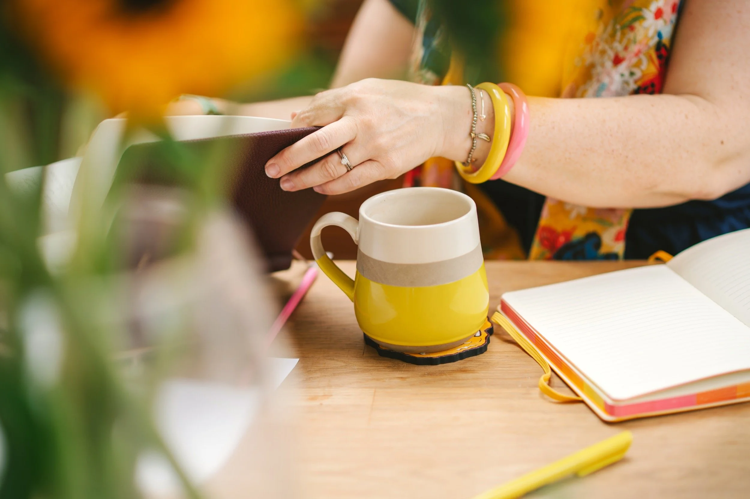 Person with colorful bracelets reading a brown notebook at a wooden table with a yellow mug, an open journal, and a yellow pen.