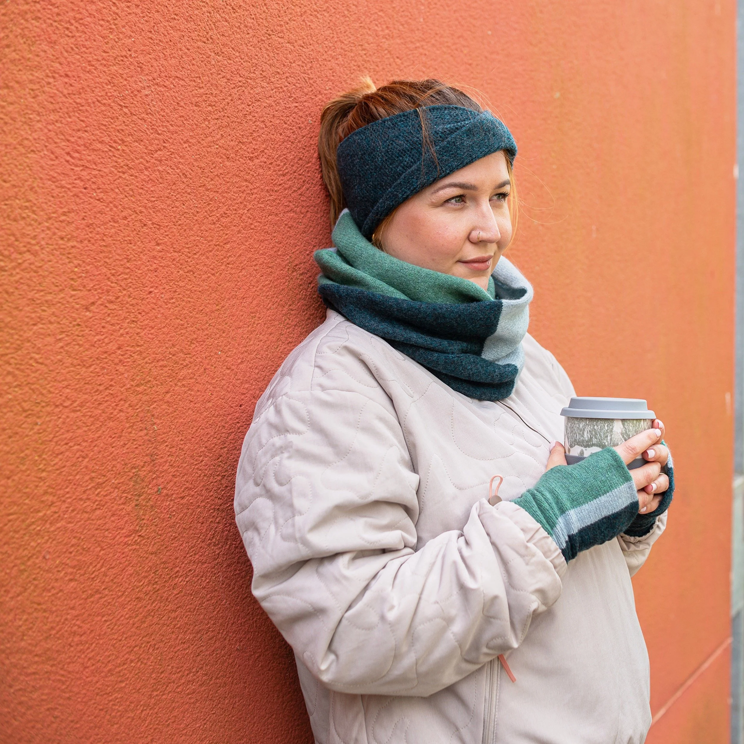 Amy leans against an orange wall, holding a travel coffee cup