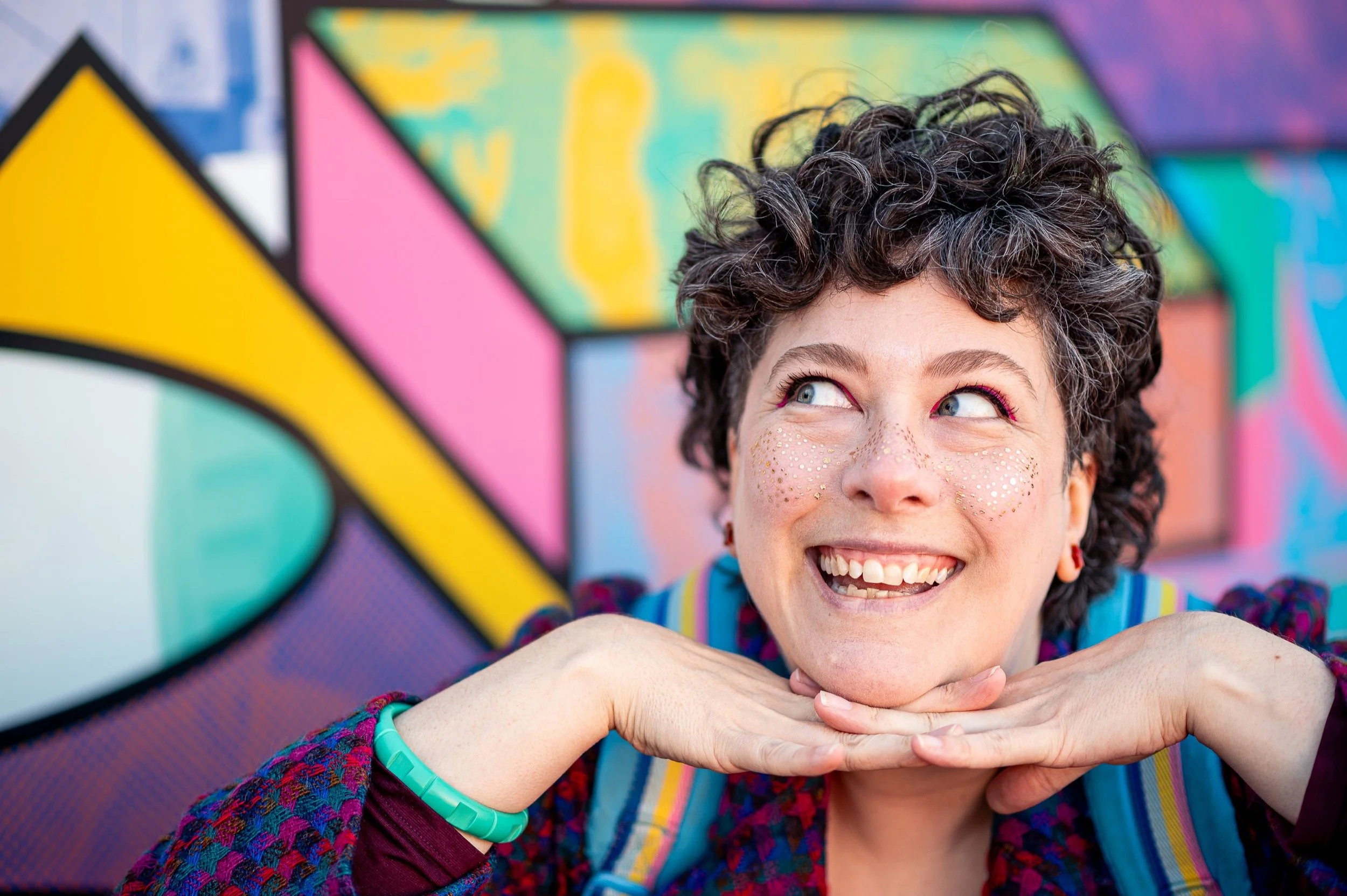 Smiling person with curly hair and glitter on their cheeks, resting their chin on her hands, in front of a colourful geometric mural.