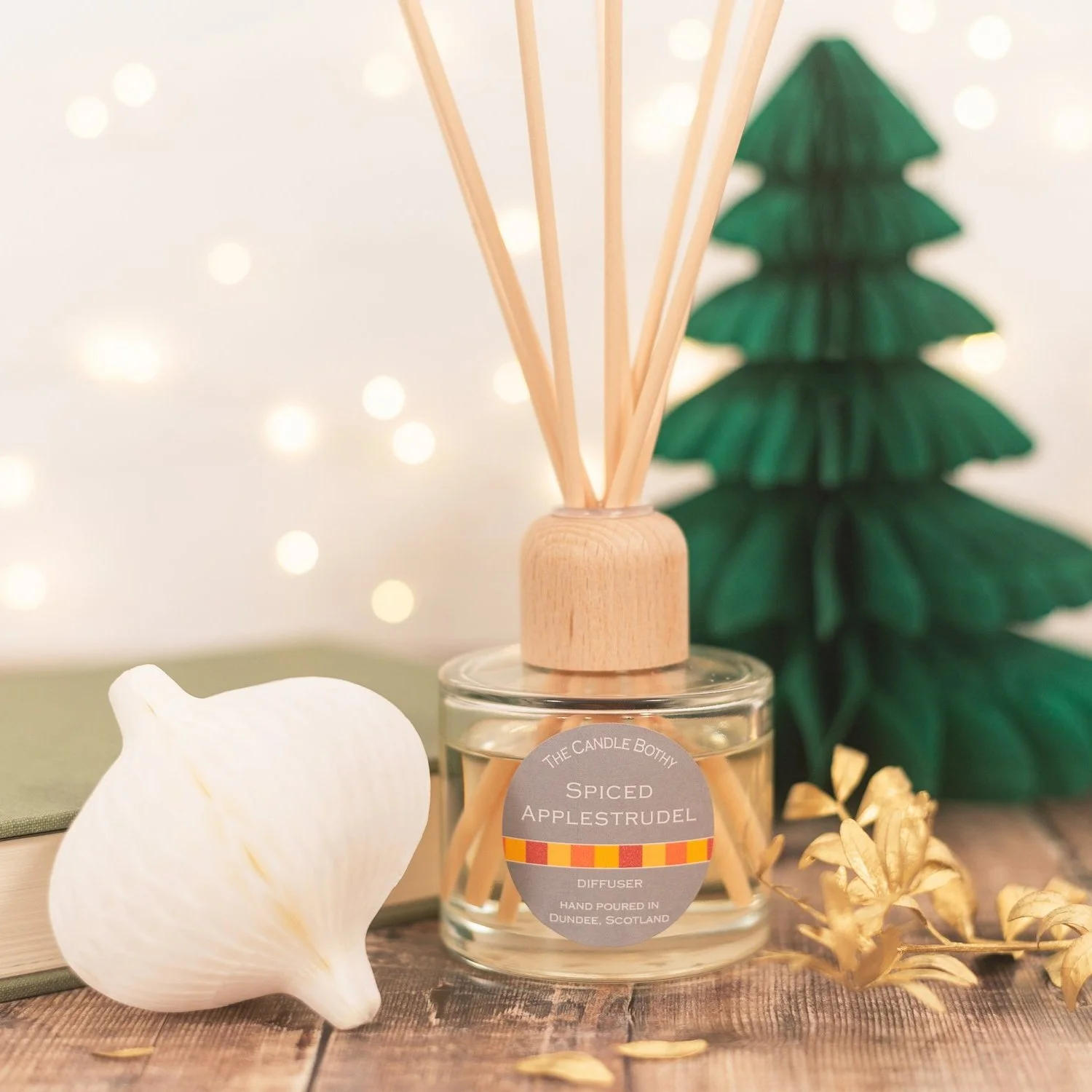 A scented reed diffuser labeled 'Spiced Appelstrudel' with beige reeds, surrounded by a white shell-shaped object, a tree-shaped green paper decoration, and gold leaves on a wooden surface with bokeh lights in the background.