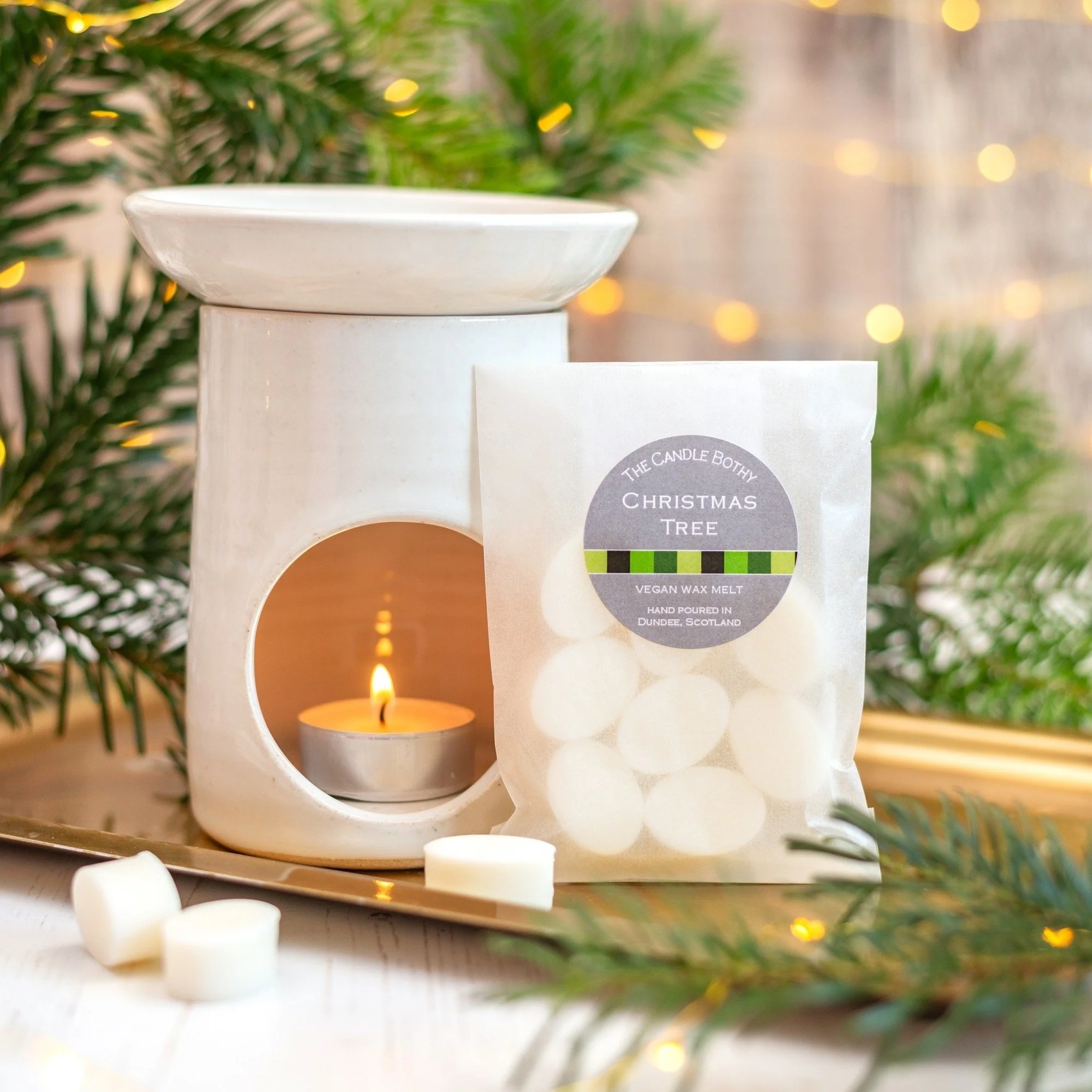 A white candle warmer with a small lit tealight candle inside, surrounded by evergreen branches and small white wax melts labeled 'Christmas Tree' placed in front of the warmer.
