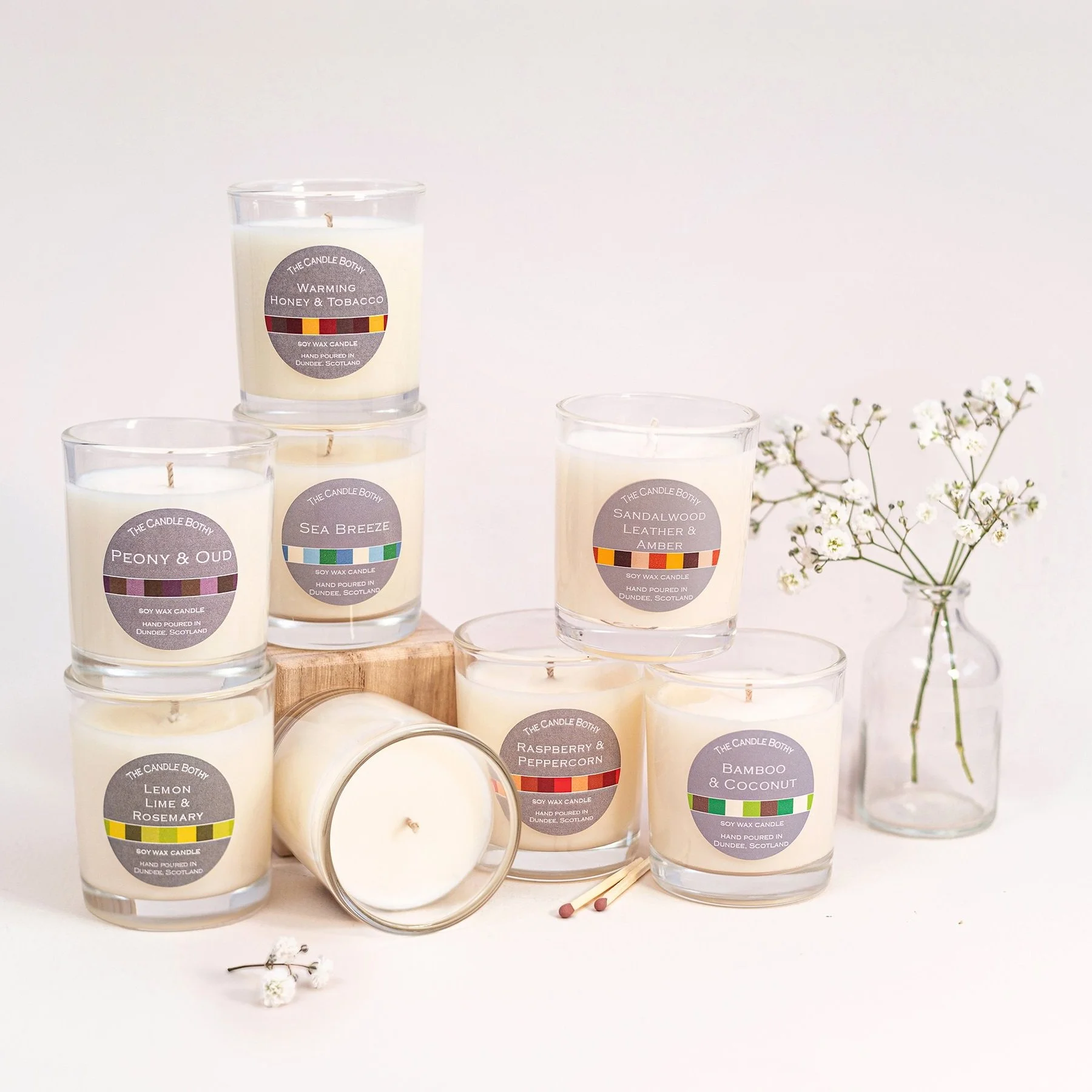 Multiple candles with floral and wooden accents. The candles have labels with scents such as 'Warming Honey & Tobacco,' 'Sea Breeze,' 'Peony & Oud,' 'Lemon Lime & Rosemary,' 'Sandalwood Leather & Amber,' 'Raspberry & Peppercorn,' and 'Bamboo & Coconu