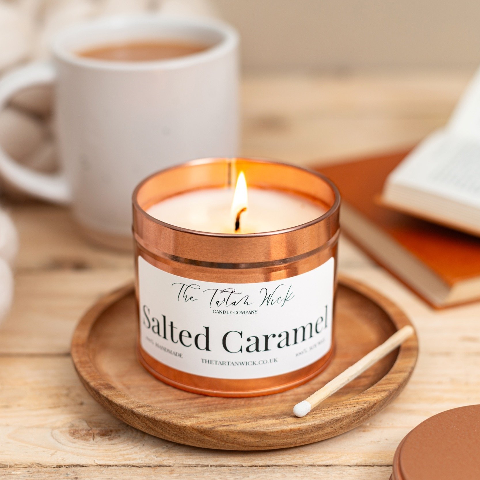 A lit salted caramel scented candle in a copper jar on a wooden tray, with a cotton swab beside it, a mug of tea, a book, and another closed container in the background on a wooden table.