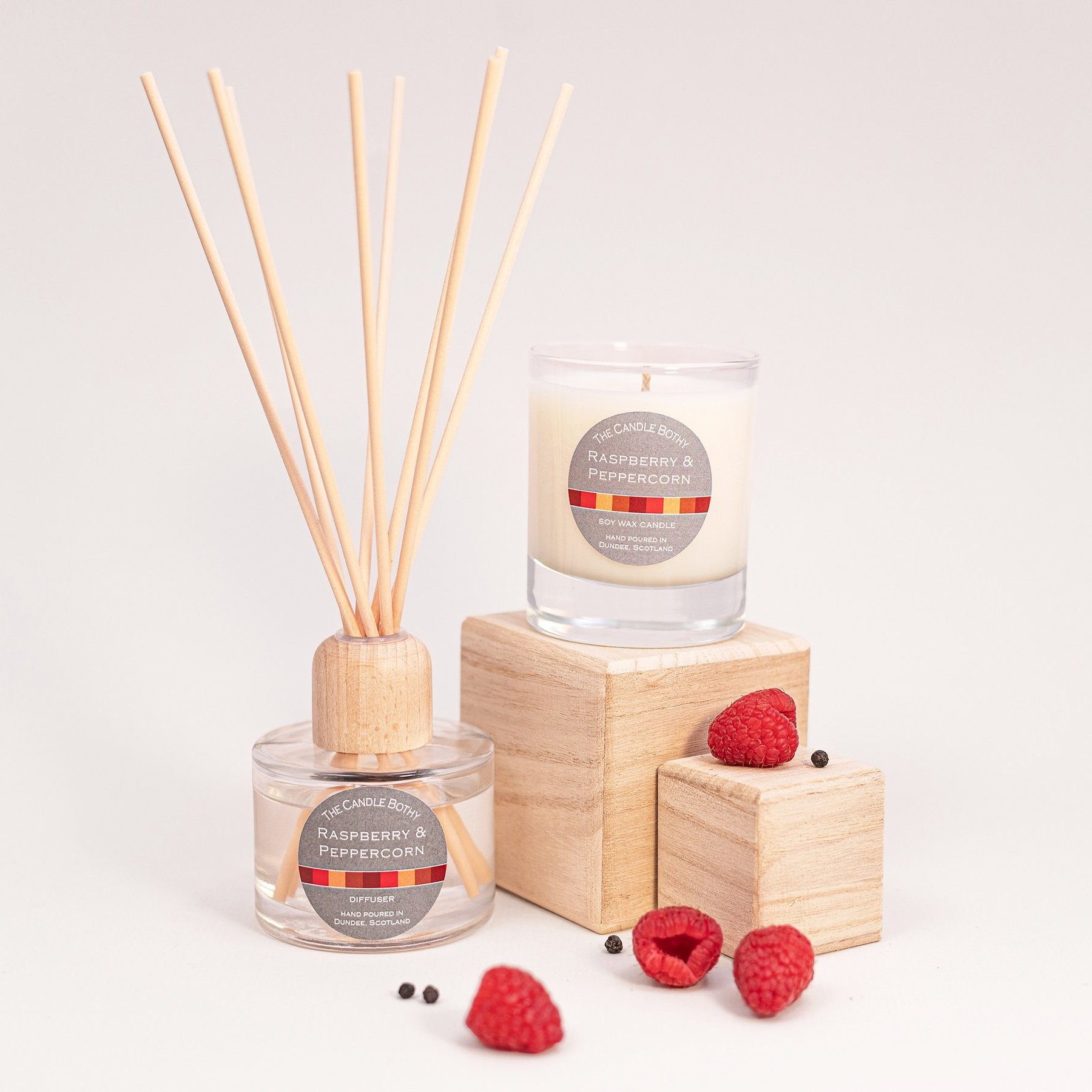 A raspberry and peppercorn scented candle, diffuser, and reed diffuser on wooden blocks with fresh raspberries and black peppercorns, against a plain white background.