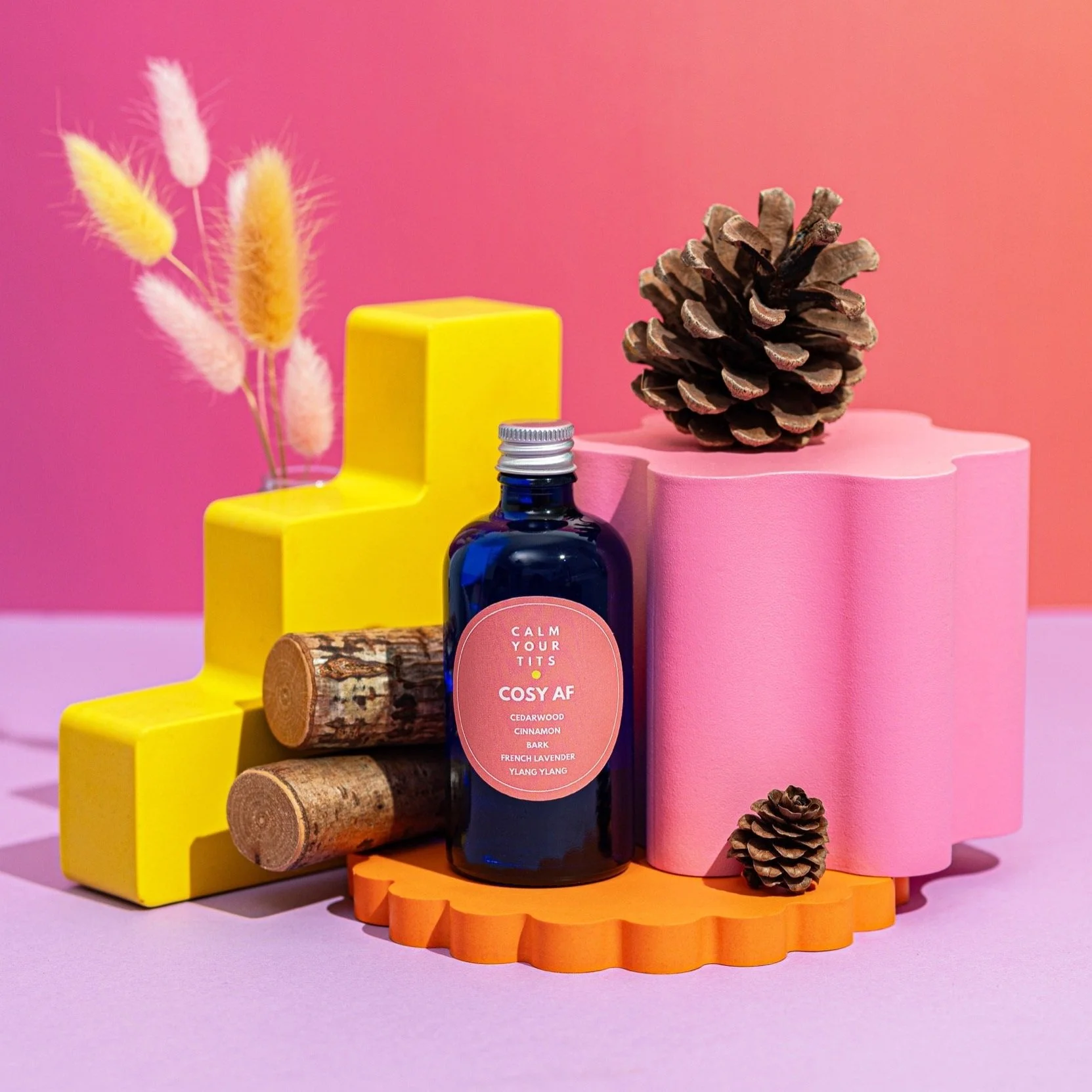 Colorful decorative arrangement with a pink spray foam block, yellow blocks, pine cones, a blue bottle labeled 'Calm Your Tits COSY AF' with cedarwood, cinnamon, bark, French lavender, and ylang ylang, and pink fuzzy plants against a pink background.