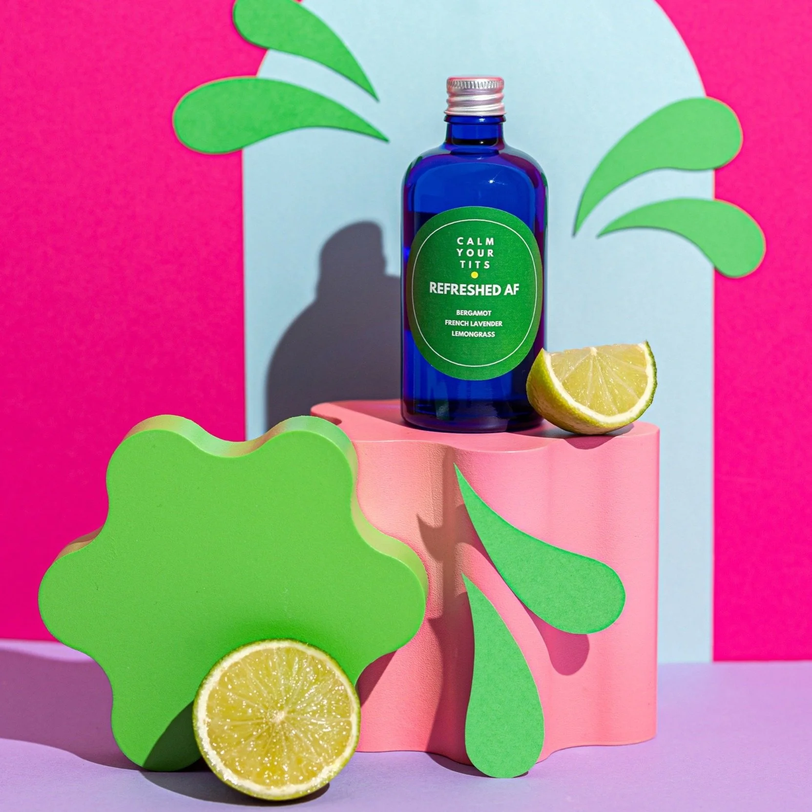 A blue bottle labeled "Calm Your Tits Refreshing AF" with bergamot, French lavender, and lemongrass, positioned on a pink block, accompanied by a lemon wedge, green paper cutouts of abstract plant leaves, and a colorful background with pink and light