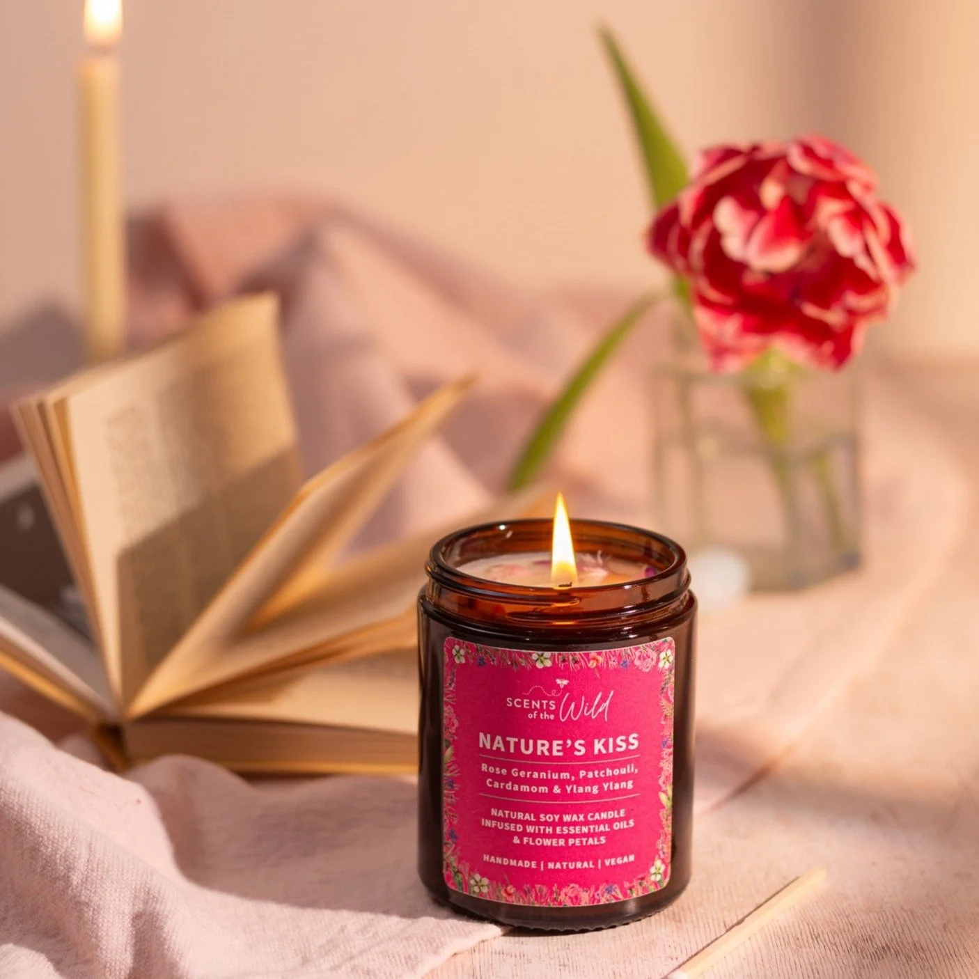 A lit candle labeled 'Nature's Kiss' with a pink floral design, surrounded by an open book, a pink cloth, and a glass vase with a pink and white flower on a soft beige surface.