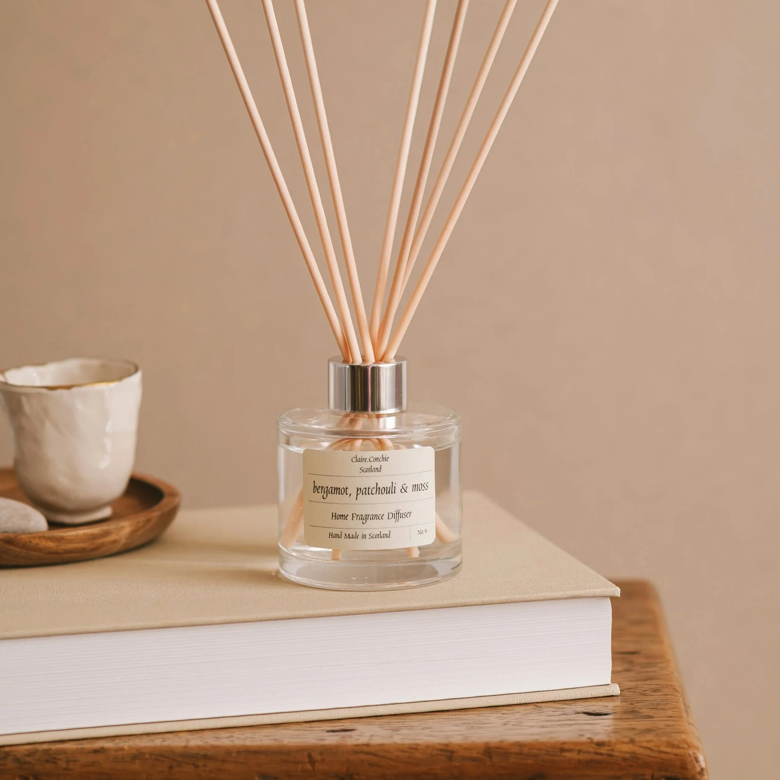 A glass jar of home fragrance diffuser with pink reed sticks, labeled 'bergamot, patchouli & moss', placed on a white book on a wooden table.