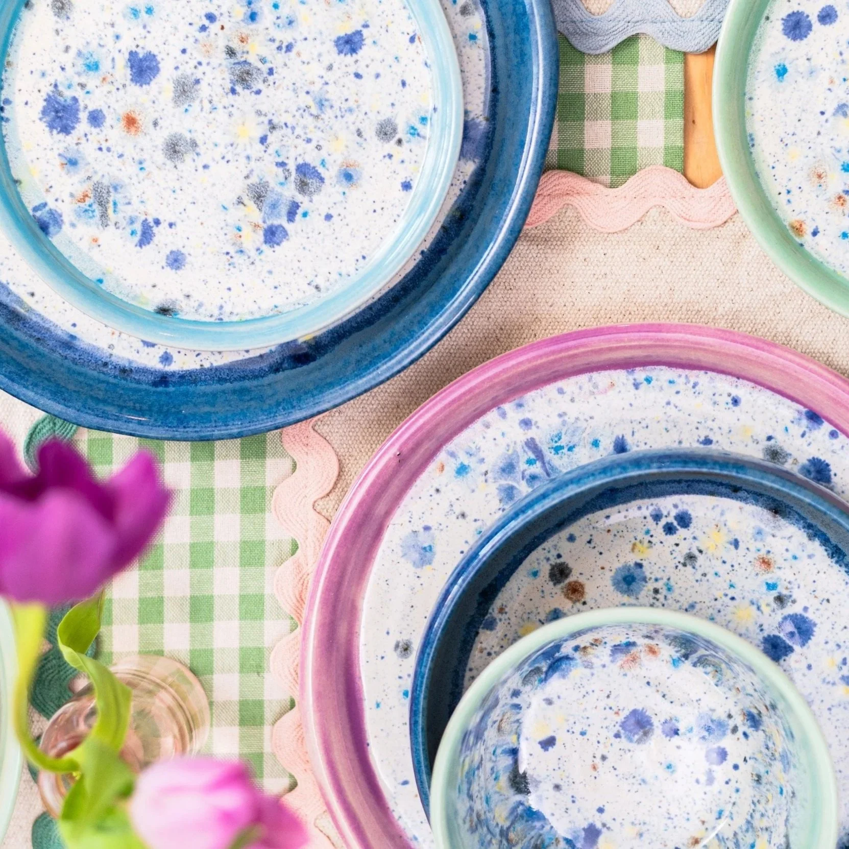 Colorful painted plates and bowls with splattered paint designs on a table with a green checkered cloth and pink lace-edged placemats, with a purple flower in the foreground.