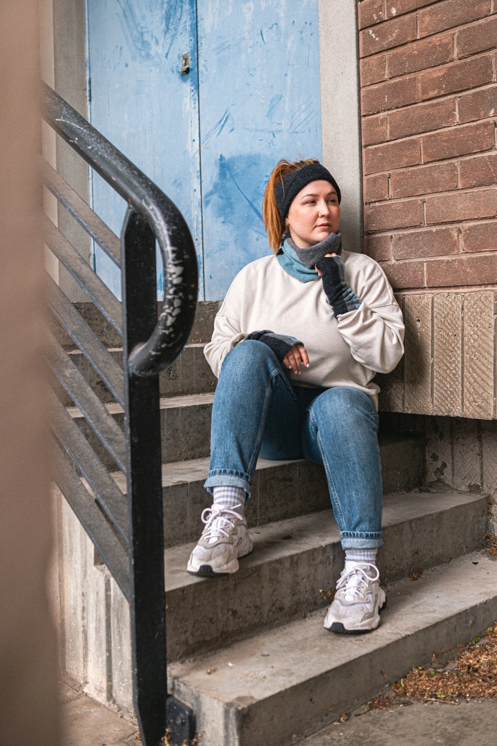 Young woman in athletic wear sitting on outdoor concrete stairs, leaning against a brick wall, wearing a black headband, gray sweatshirt, blue jeans, and sneakers, with a multicoloured neck warmer and gloves, looking thoughtfully to the side.