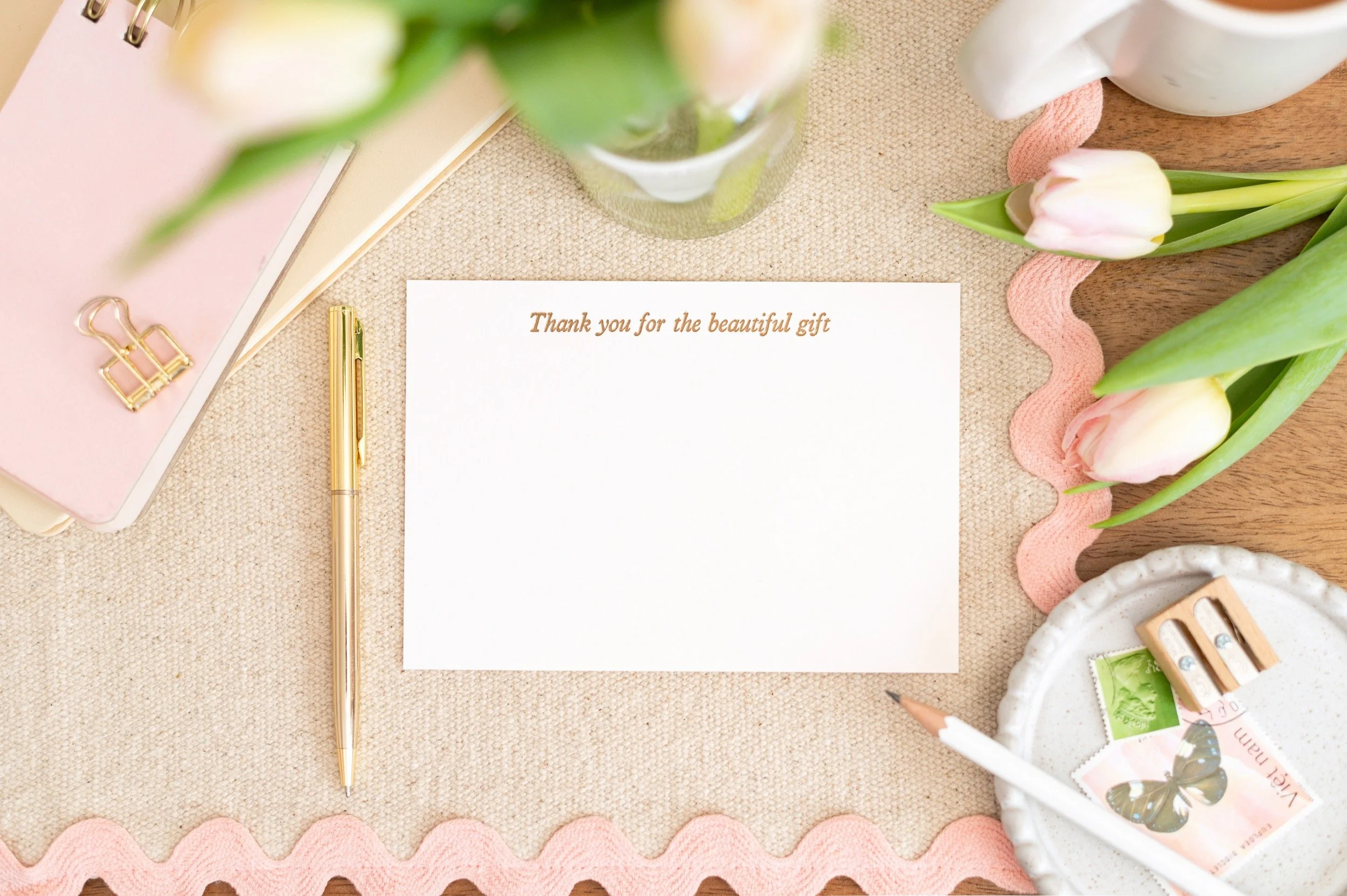 Flat lay of a thank you card on a beige cloth, surrounded by pink tulips, a white mug, a gold pen, a pink notebook, a small white dish with postage stamps, a pencil, and a pink rickrack trim.