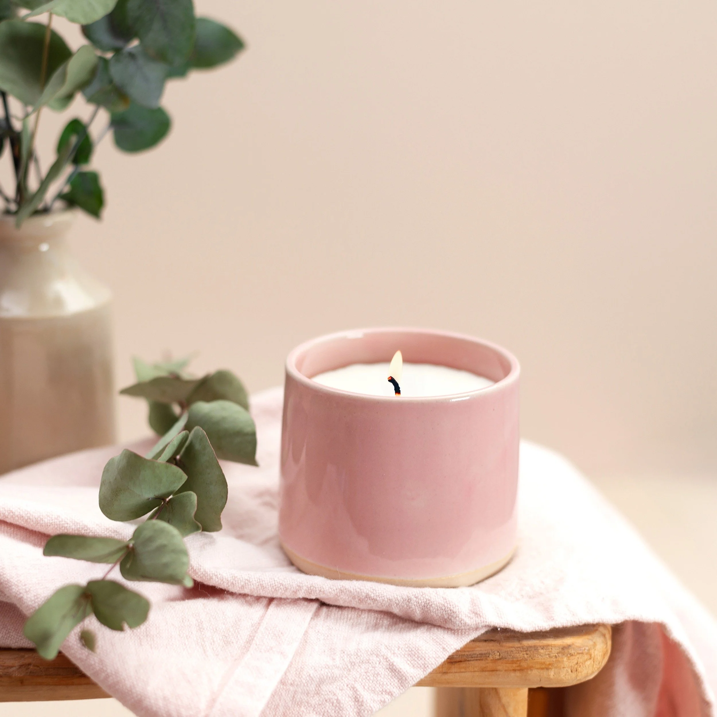 Pink ceramic candle holder with lit candle, green eucalyptus leaves on a pink cloth, and a beige vase with eucalyptus in the background.