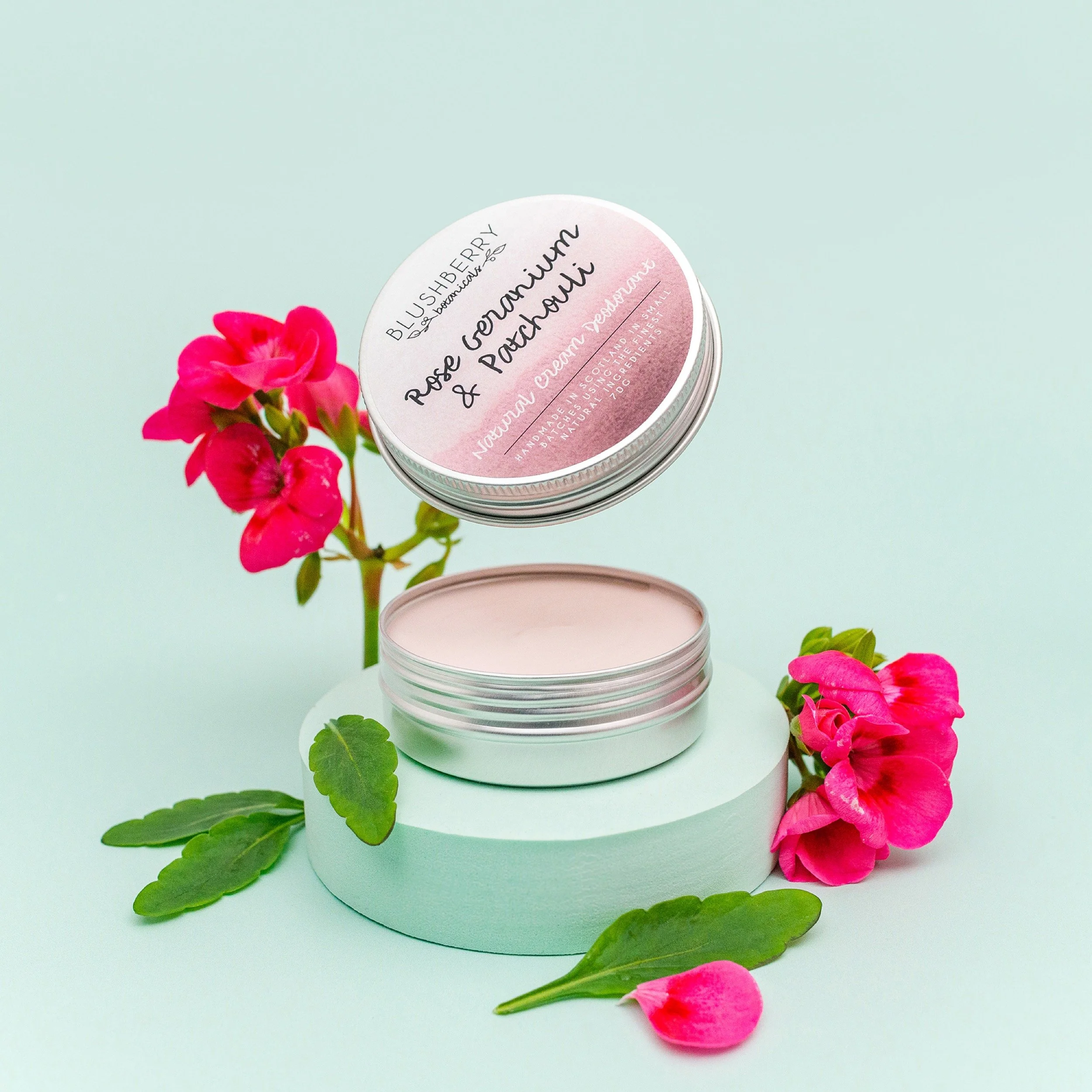 Open tin of Rose Geranium and Patchouli hand cream surrounded by pink flowers and green leaves on a pastel background.