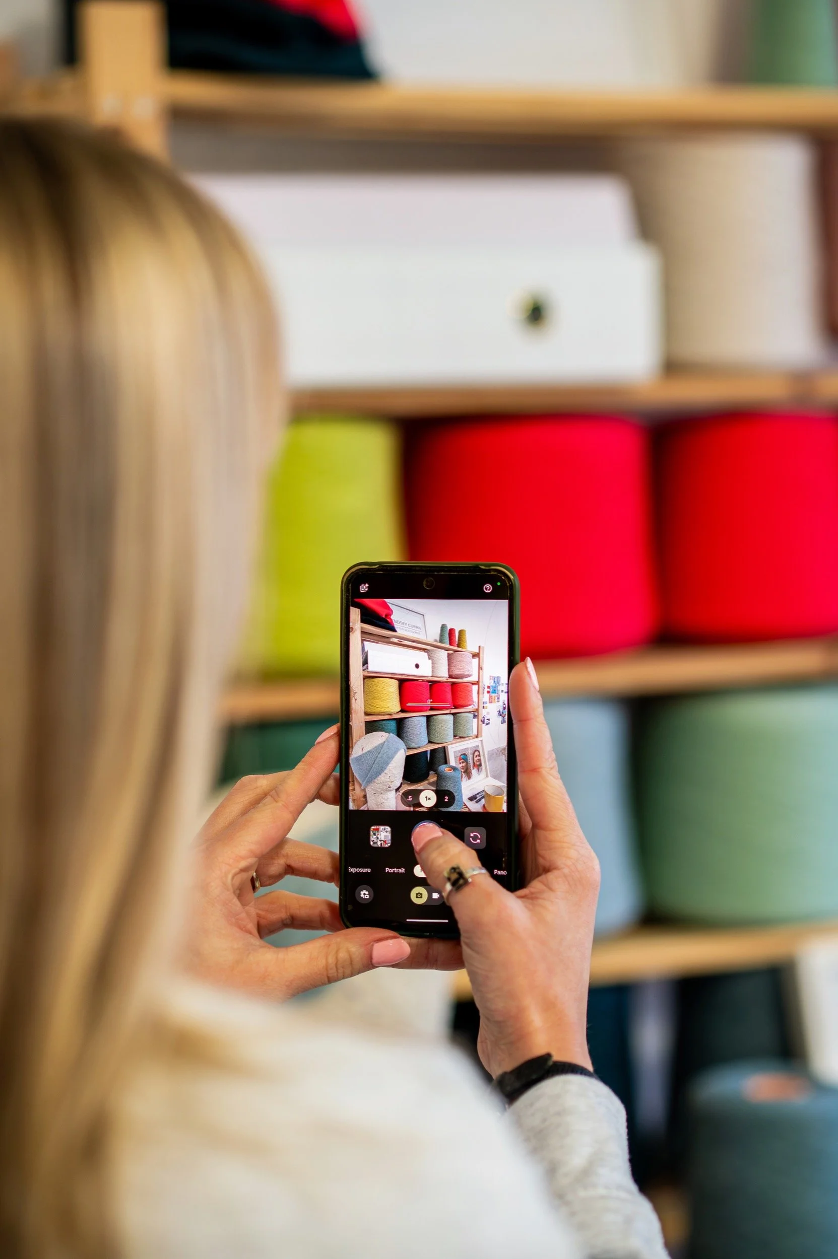 Person taking a photo of a colourful wool display on a wooden shelf with a smartphone.