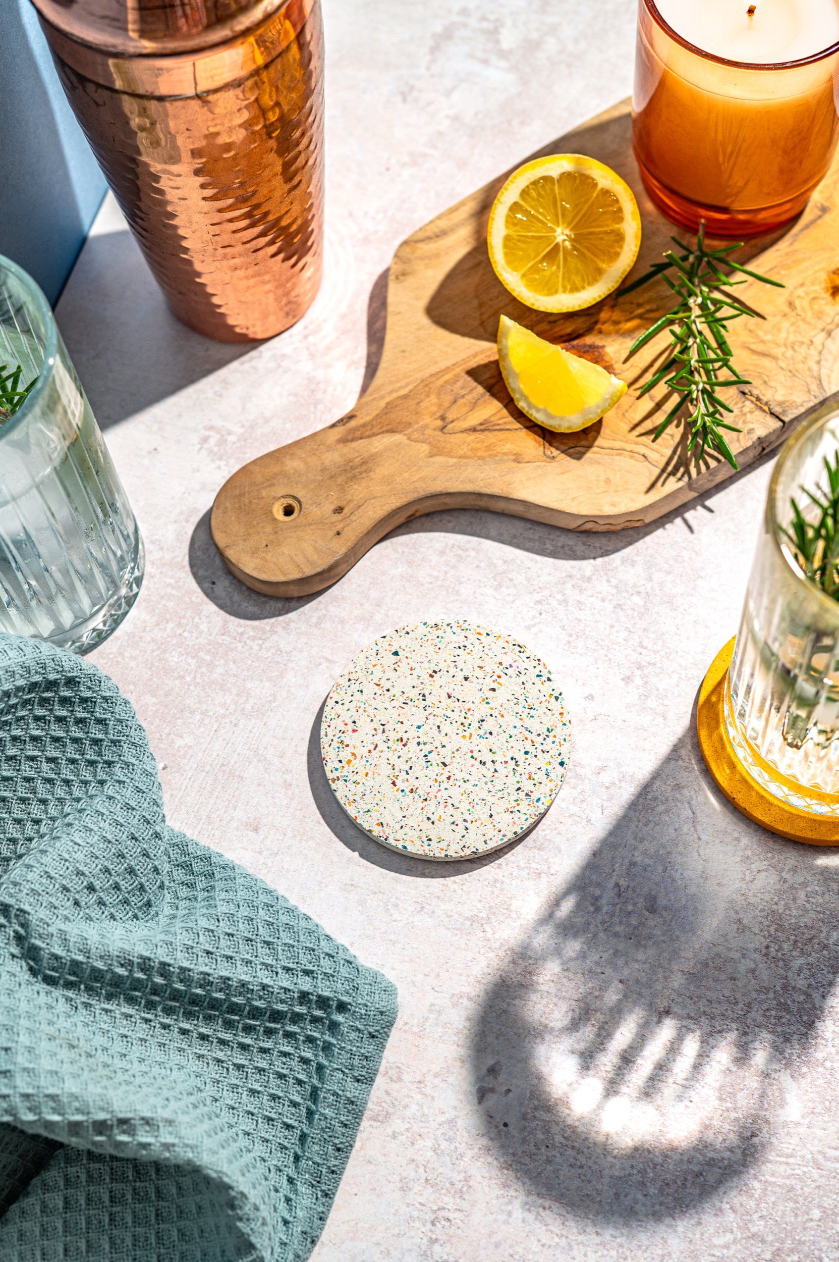 a terrazzo effect jesmonite coaster sits on a worktop with a chopping board and cocktail garnishes