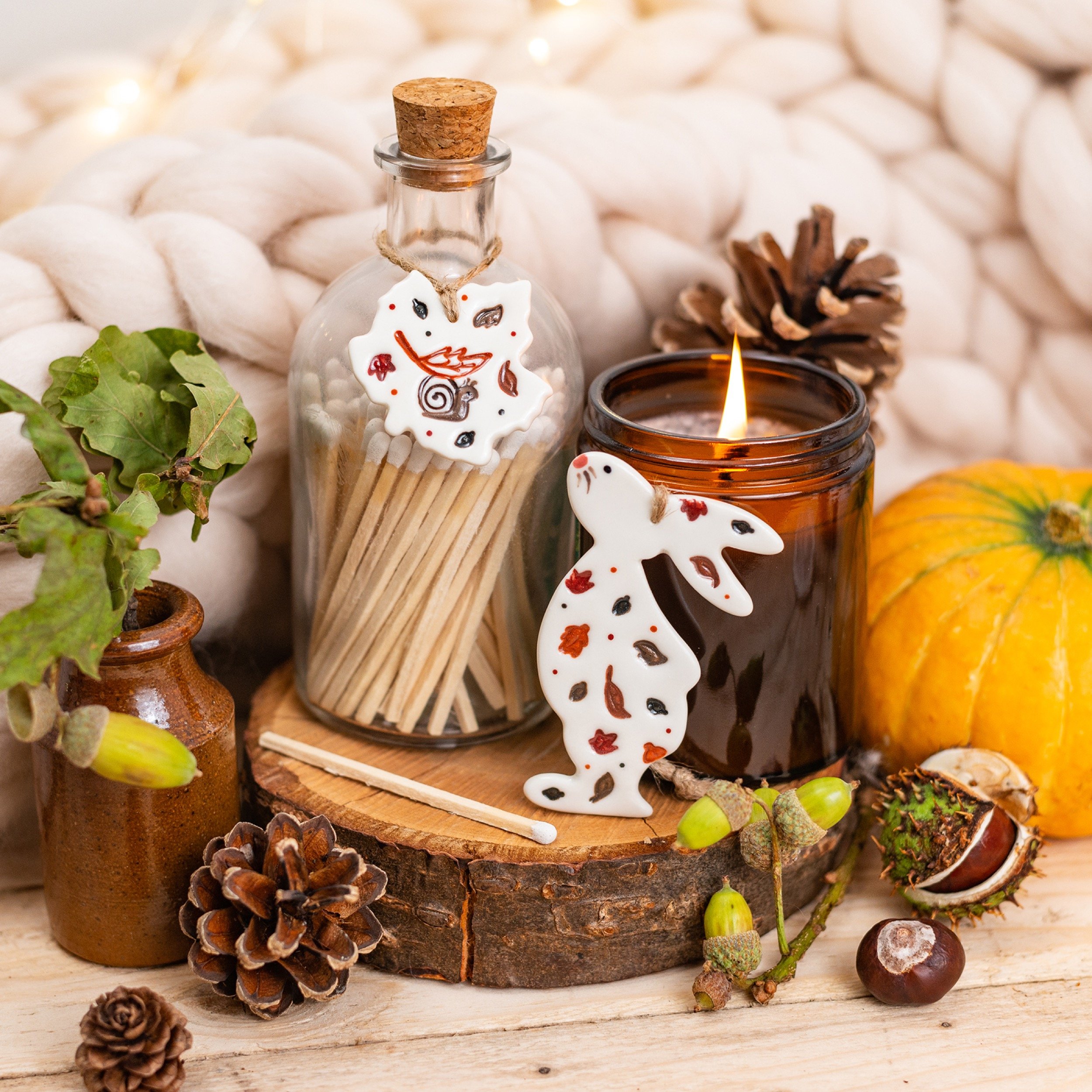 Autumn-themed decor featuring a small lit candle in a brown glass jar, a glass jar filled with toothpicks with a leaf-shaped porcelain ornament, pumpkins, pinecones, acorns, a small brown bottle, and a white plush blanket in the background.