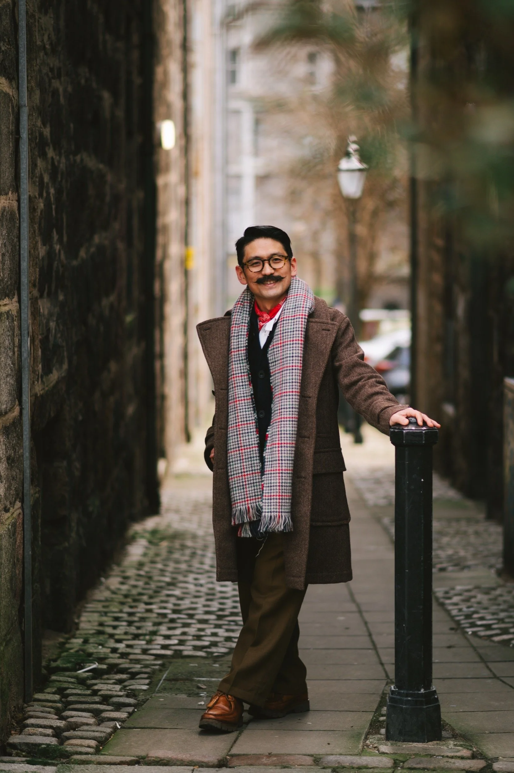 A man with glasses, a mustache, and dark hair stands on a cobblestone street, smiling, wearing a brown overcoat, a checkered scarf, and brown trousers.