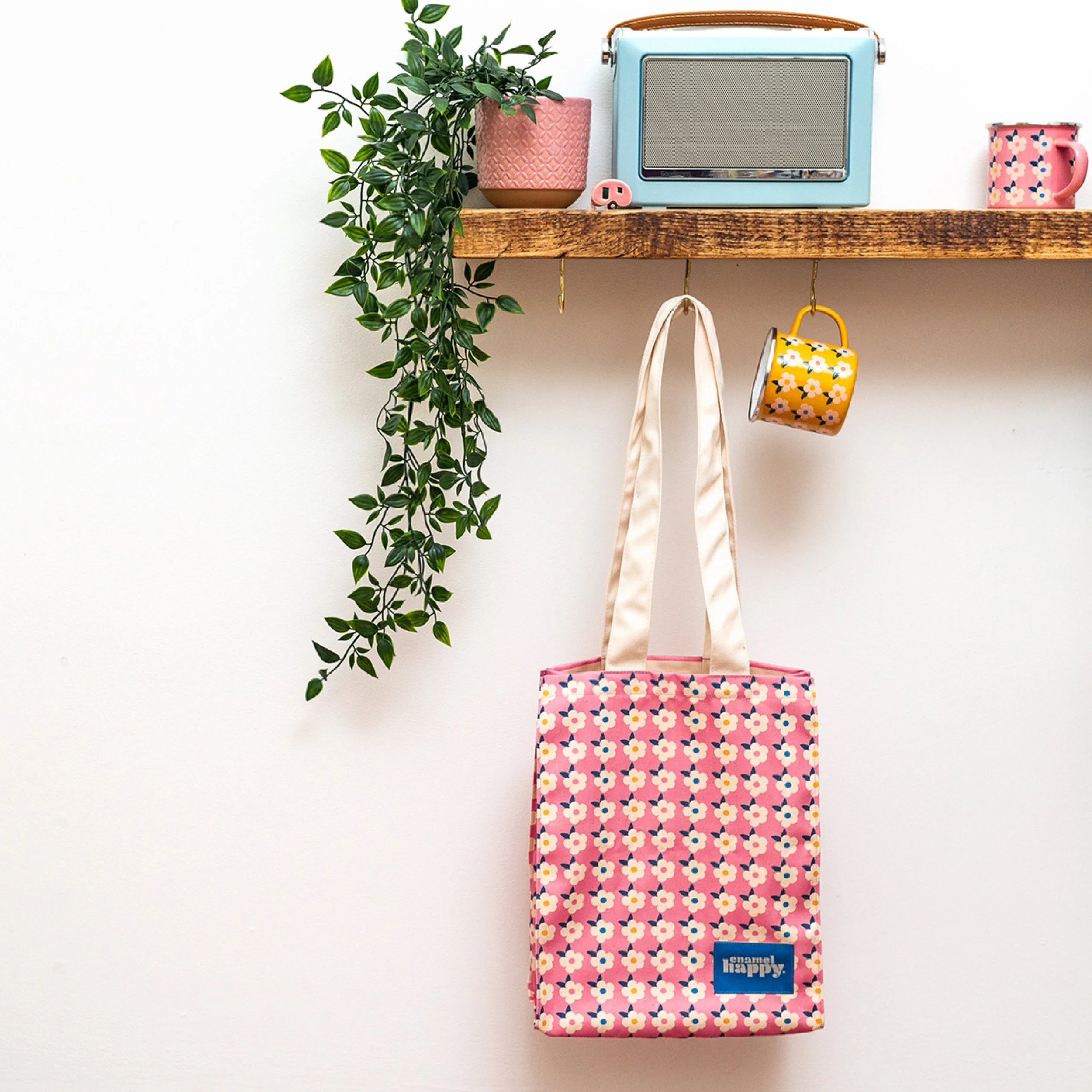 Decorative wall shelf with potted plant, vintage radio, floral mug, hanging tote bag with pink flowers, and a floral cup on a white wall.