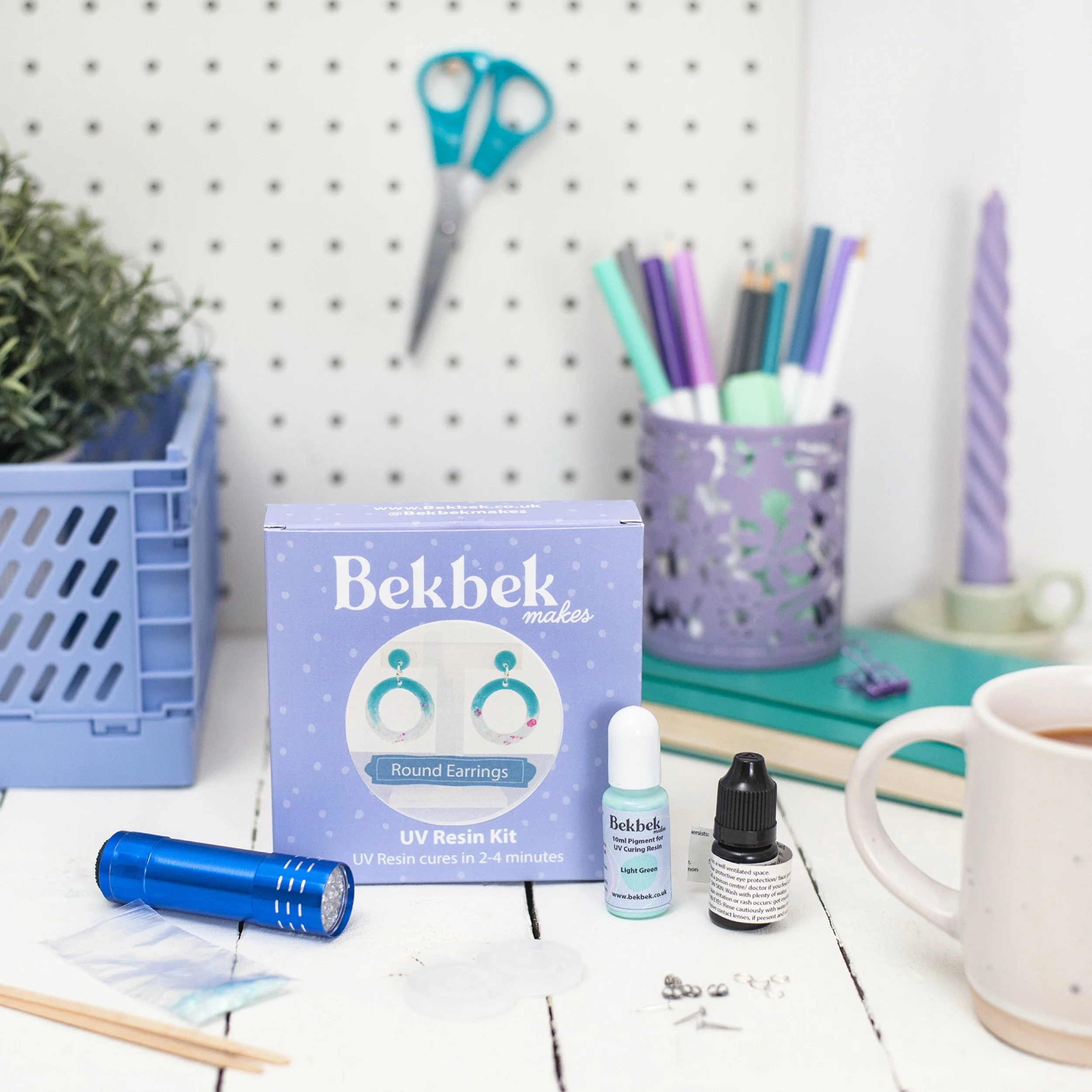 Desk setup with a Bekbek UV resin kit for making round earrings, UV flashlight, bottles of UV curing resin and pigment, jewelry findings, and various office supplies including scissors, pens, and a cup of tea.
