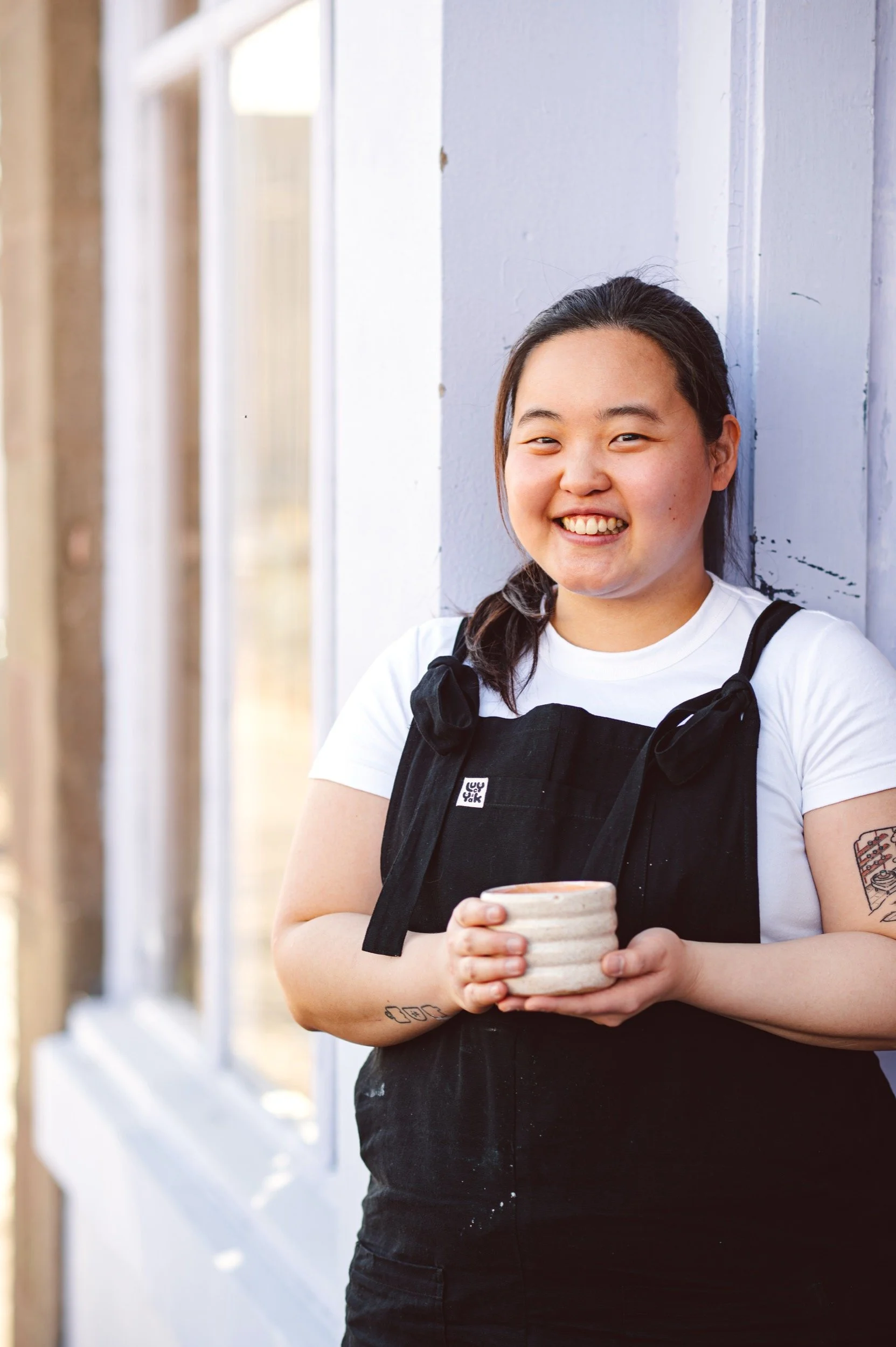 A young woman smiling and holding a ceramic cup, standing outdoors against a light-coloured wall.