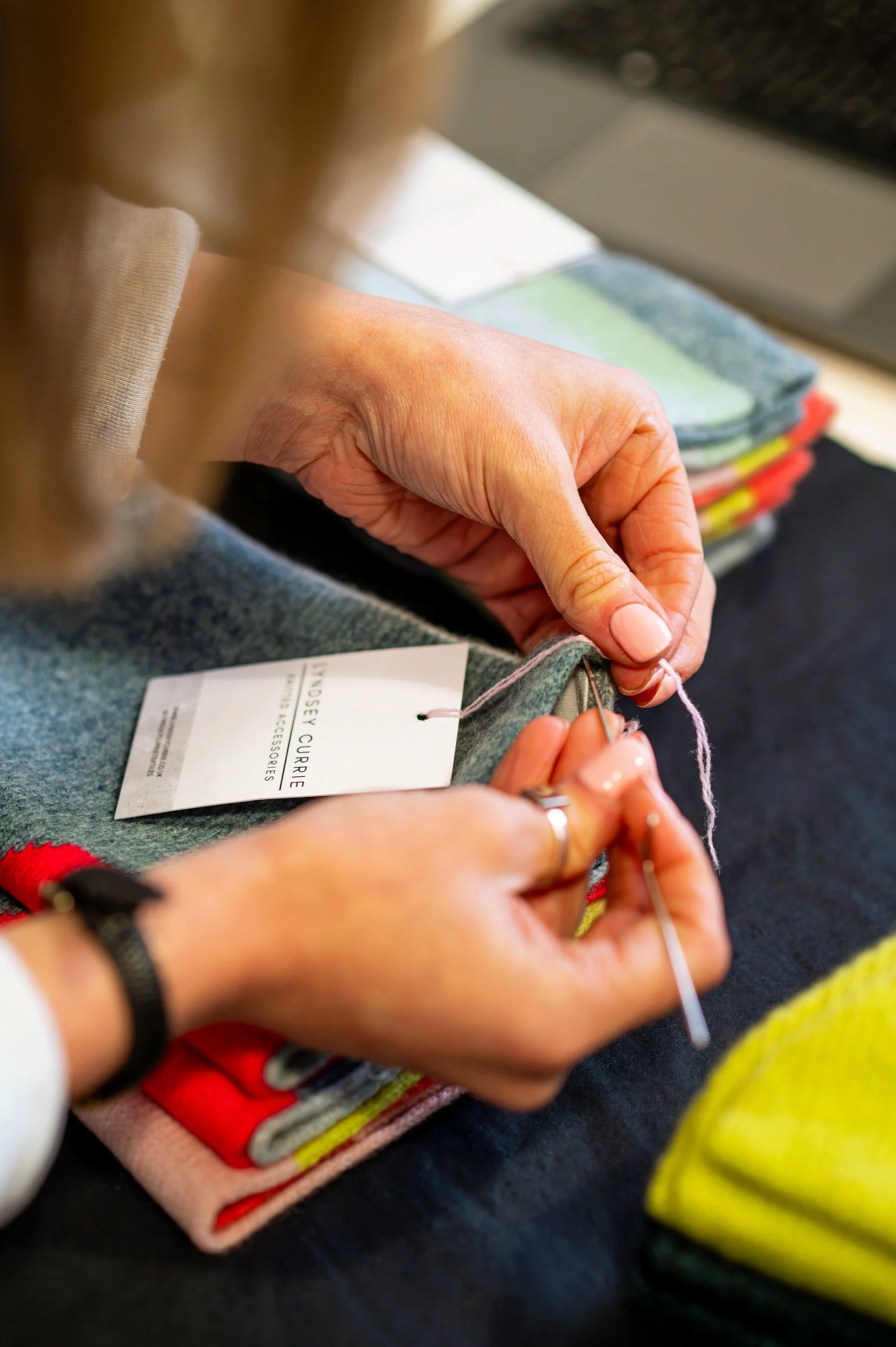 Close-up of hands sewing gray fabric with a needle and pink thread, with folded colorful fabrics in the background.
