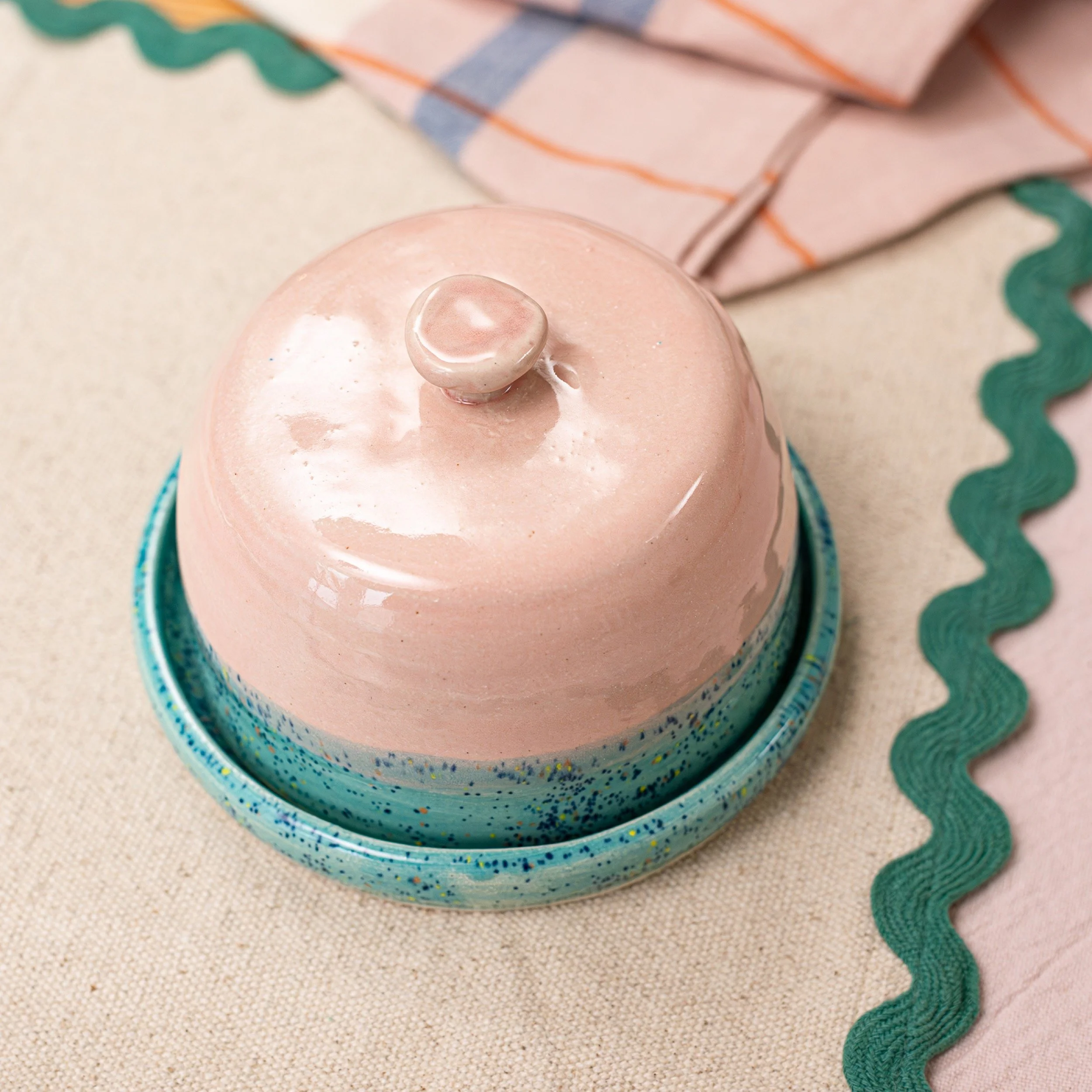 A pale pink ceramic cupcake-shaped container with a shiny finish, sitting on a speckled blue ceramic plate, with a beige tablecloth underneath and colorful paper decorations nearby.