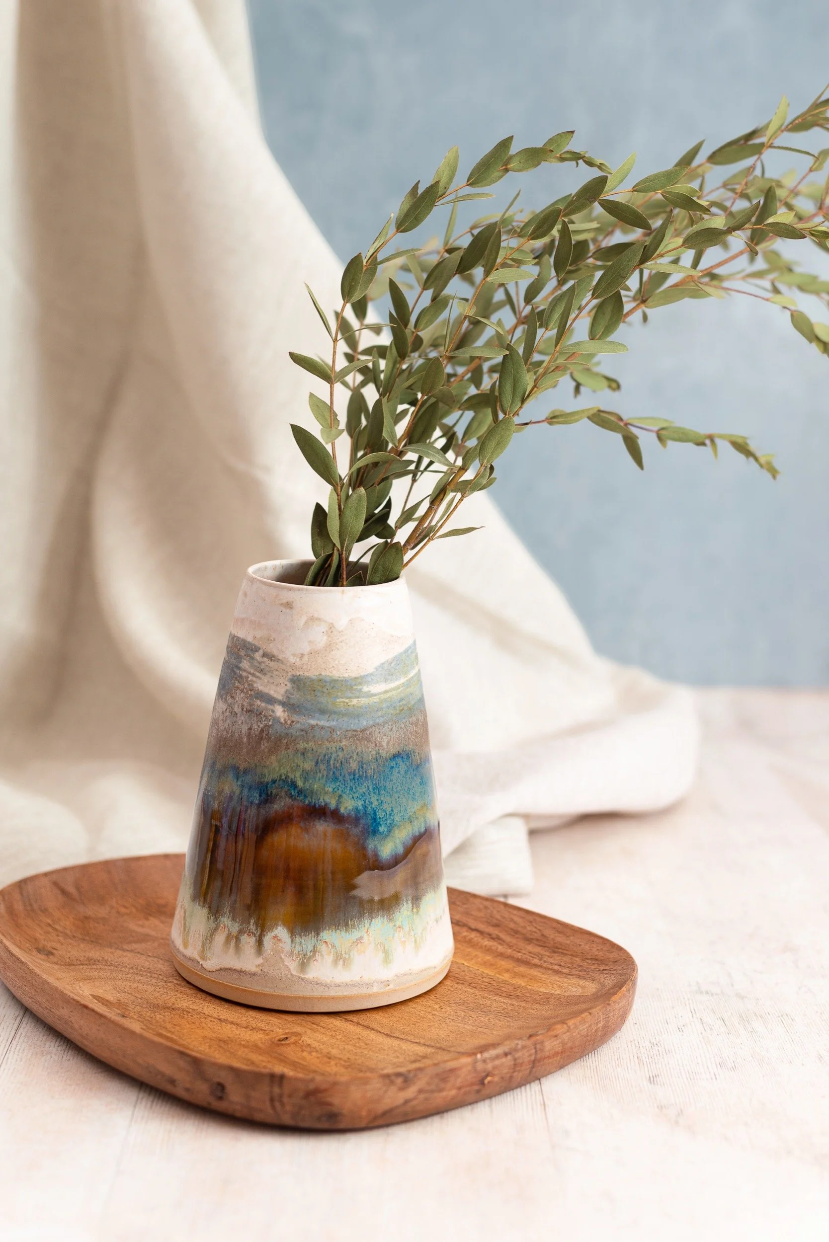 A ceramic vase with a watercolor-style design in earth tones and blues, containing green leafy branches, placed on a wooden tray with a beige fabric background and light blue backdrop.