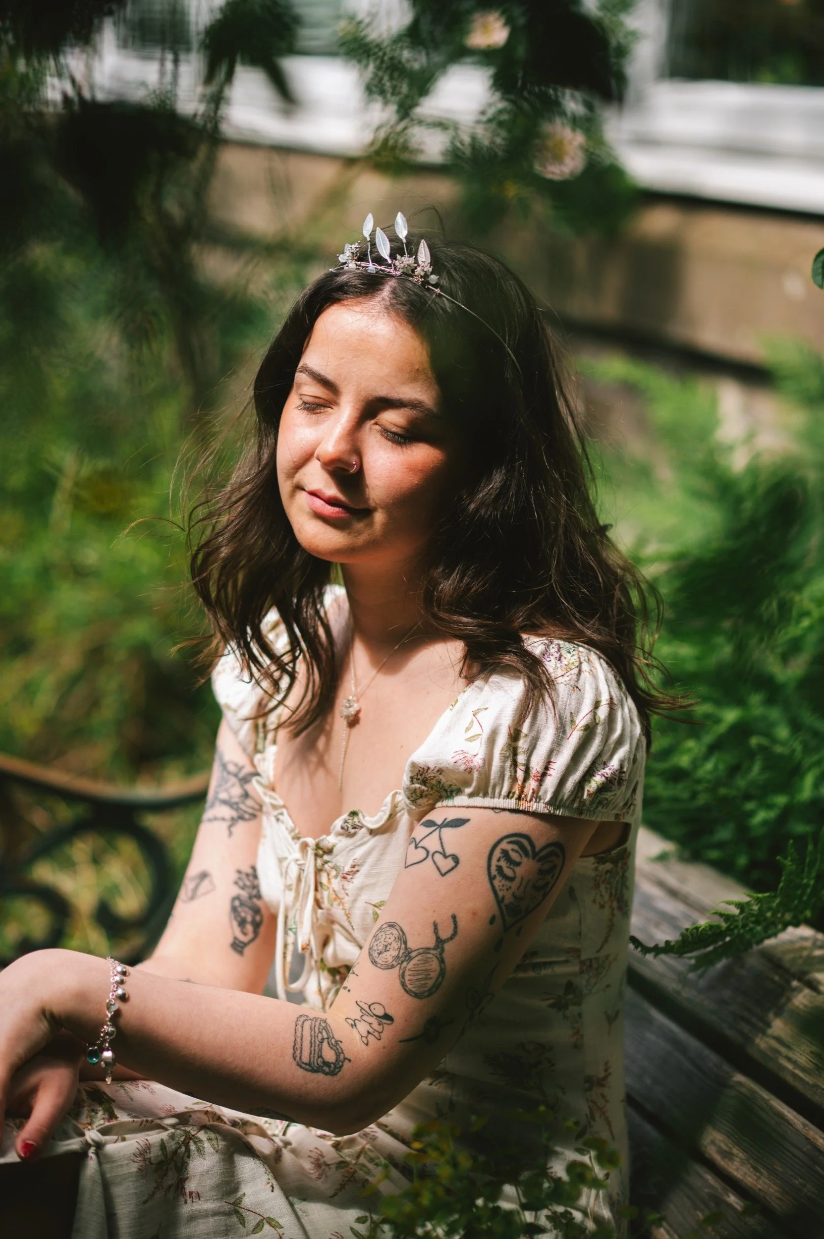 A young woman with dark wavy hair wearing a floral dress and a silver necklace, sitting outdoors among greenery. She has tattoos on her arms, and is wearing a silver bracelet. She has a small silver tiara on her head and is looking down with her eyes