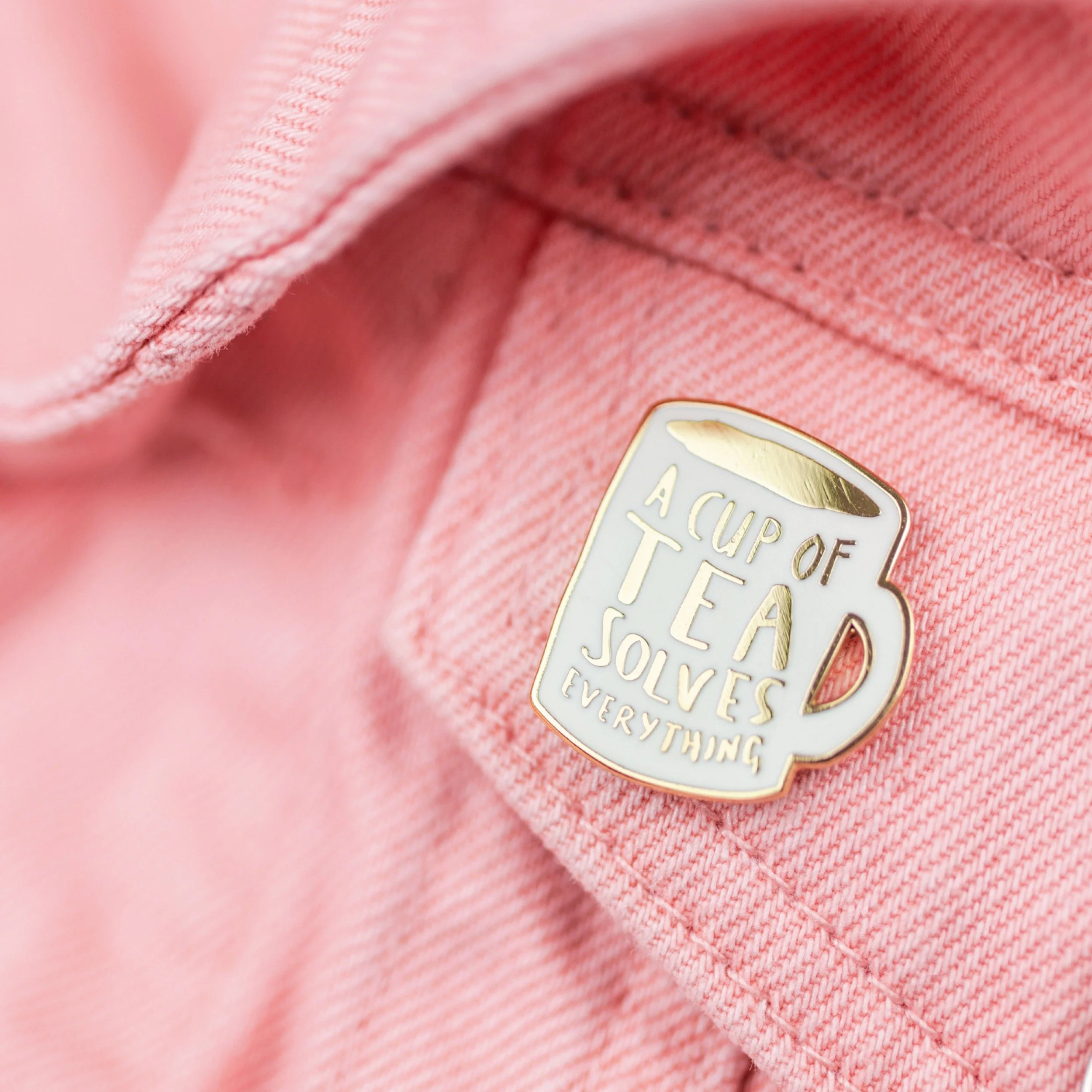 Pink denim jacket with a metal pin shaped like a mug that reads, "A cup of tea solves everything."