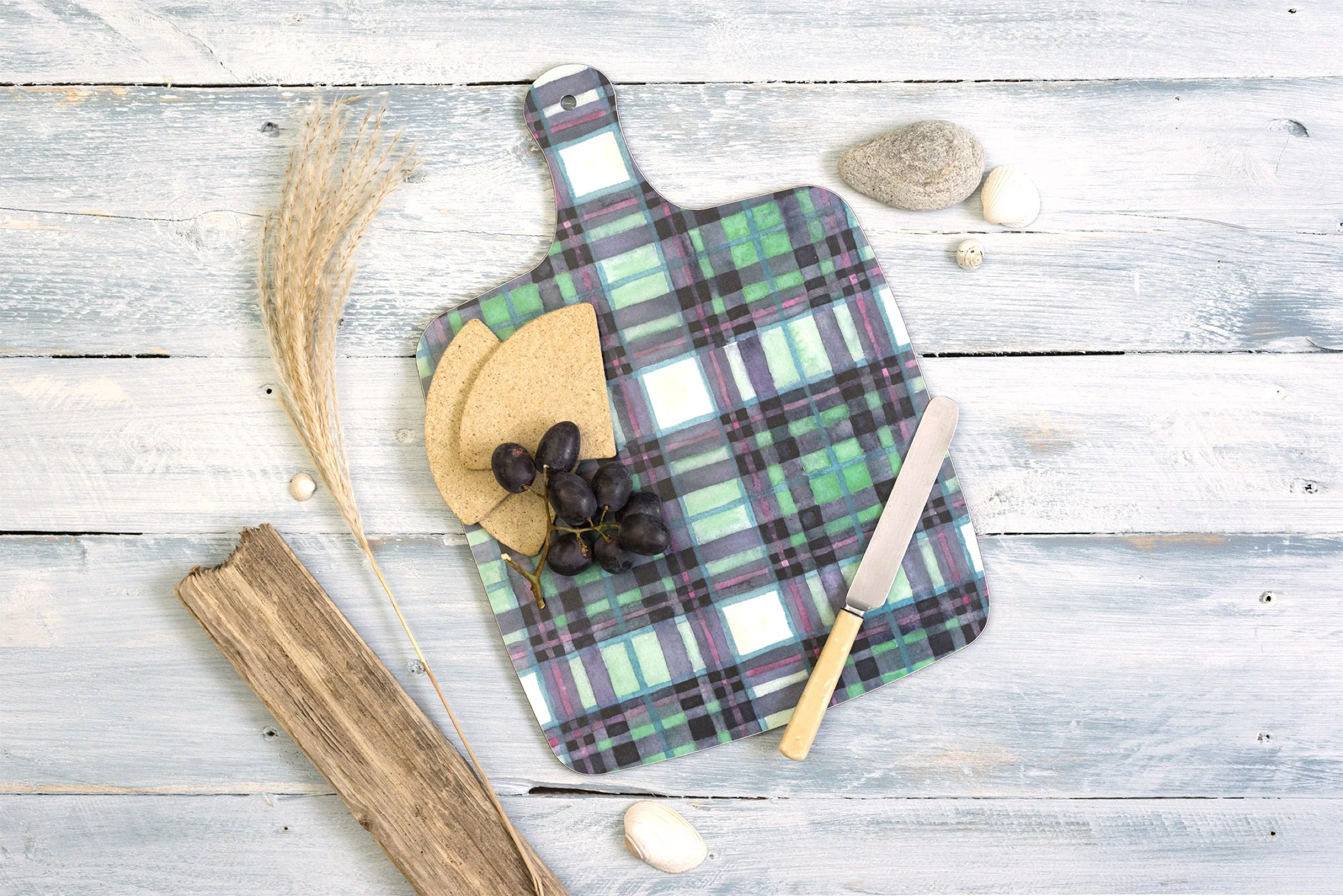 A decorative cutting board with a plaid pattern, several crackers, a bunch of black grapes, and a cheese knife, on a rustic wooden surface with rocks and seashells around.