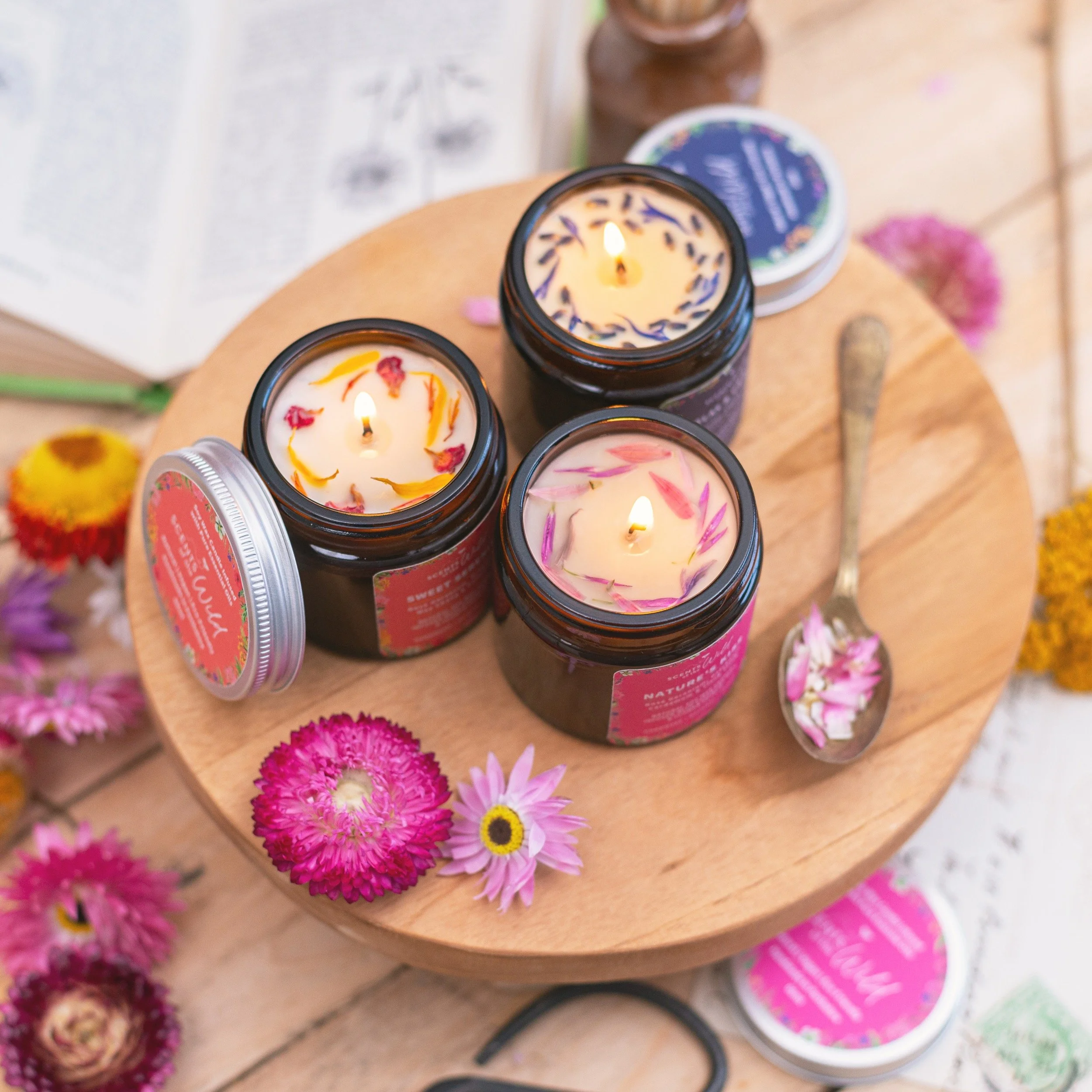 Three lit scented candles in brown glass jars placed on a round wooden tray, with a metal spoon containing flower petals nearby. Vibrant pink, purple, and yellow flowers are scattered around the tray, and an open book is in the background.