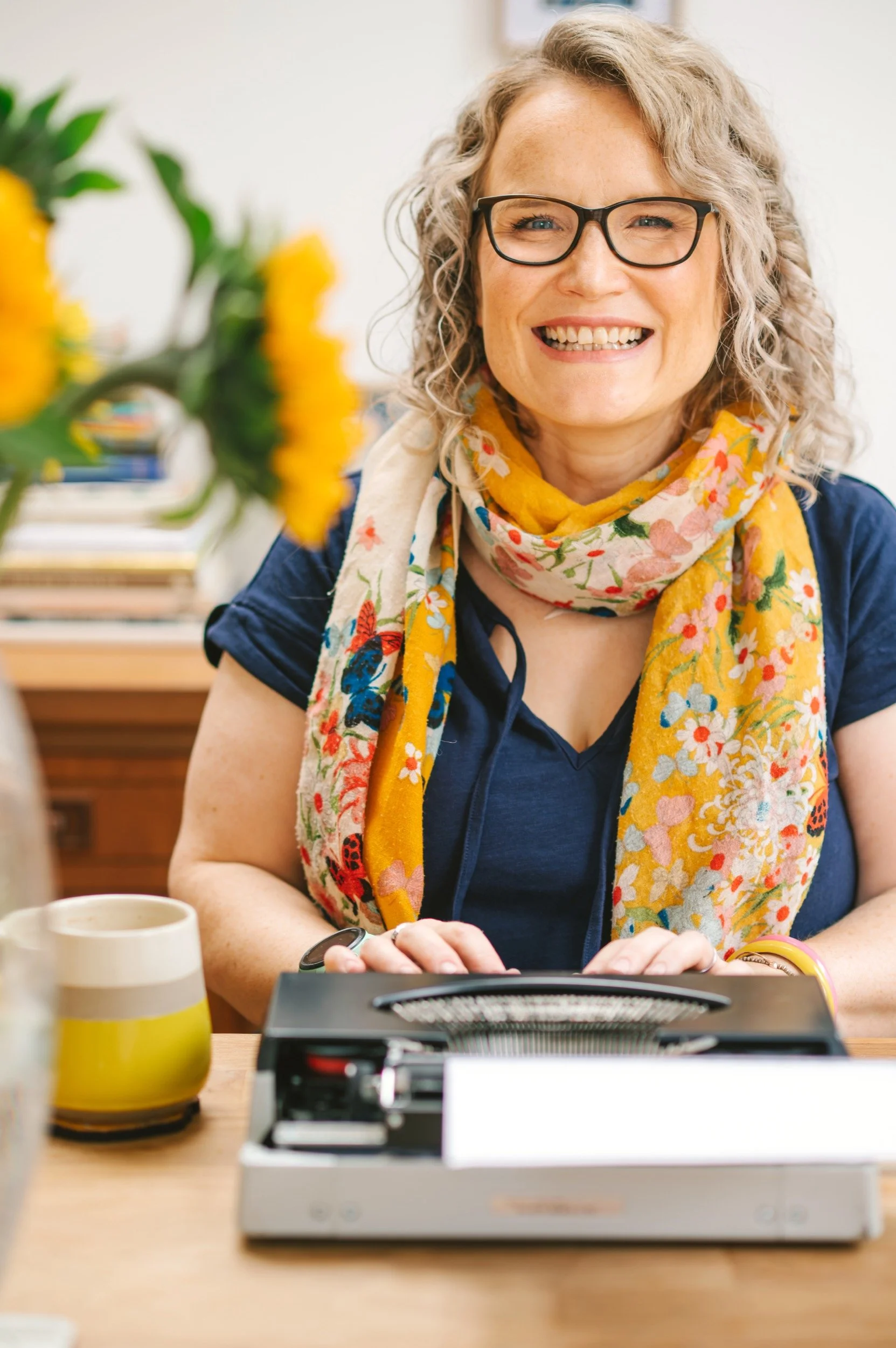 A smiling woman with curly blonde hair, glasses, and wearing a colorful floral scarf sitting at a desk with a vintage typewriter, a yellow and white mug, and out of focus flowers in the foreground.