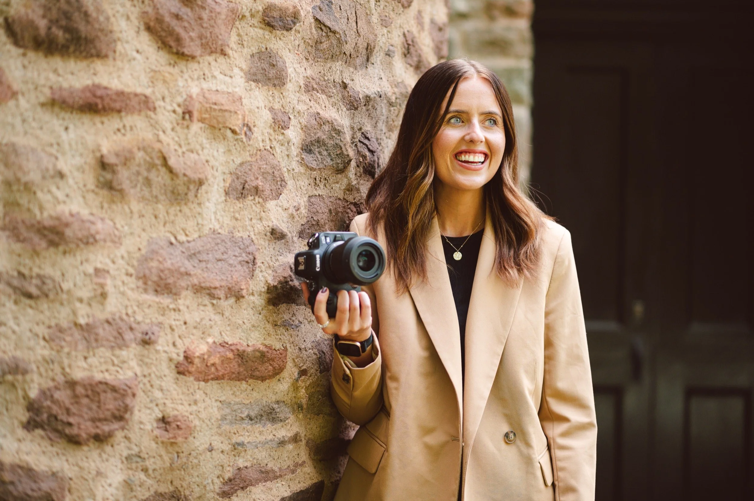 Lauren stands next to a sandstone wall which matches her camel coloured coat, she's holding her camera