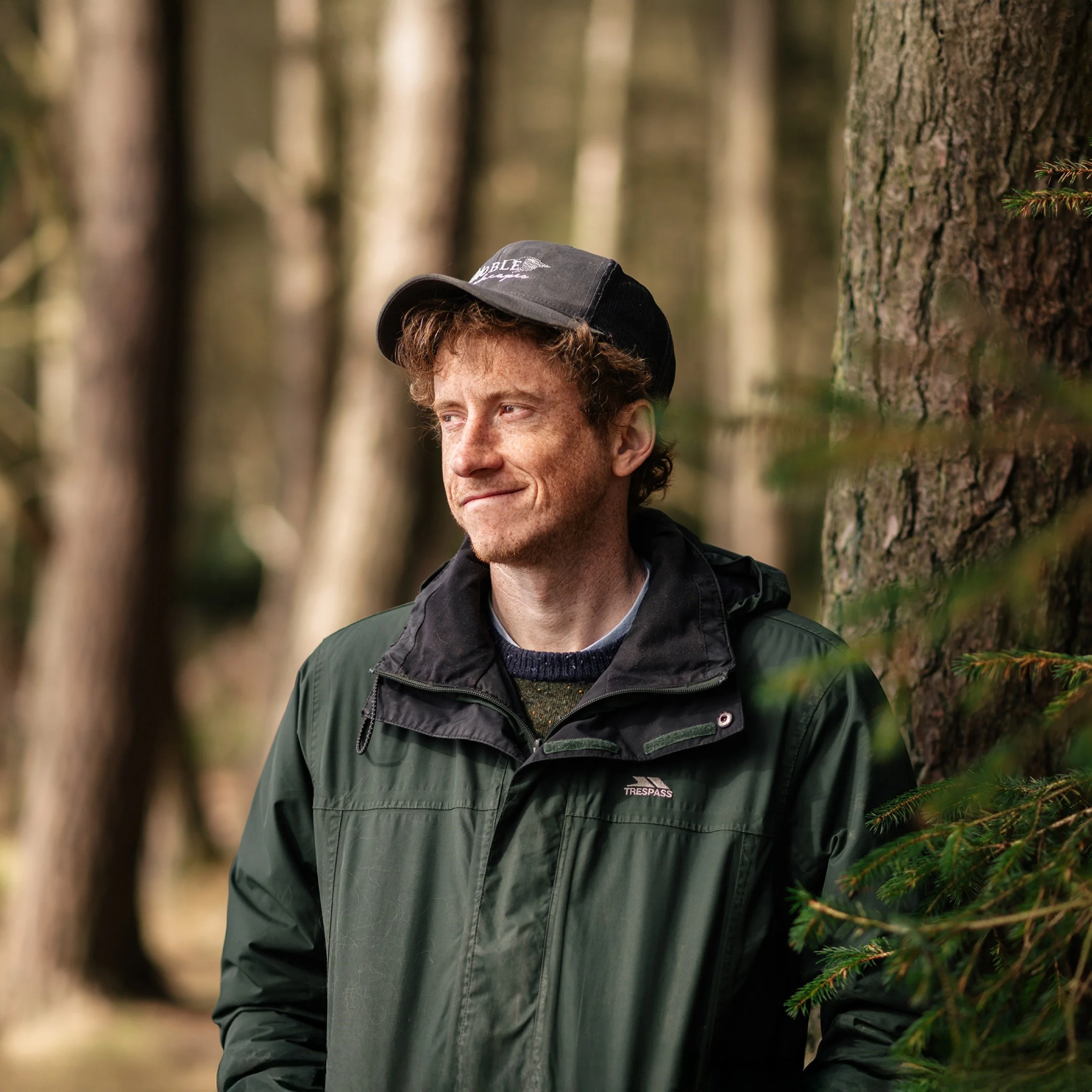 A man with reddish hair, wearing a grey cap and a green jacket, standing in a forested area with trees in the background.