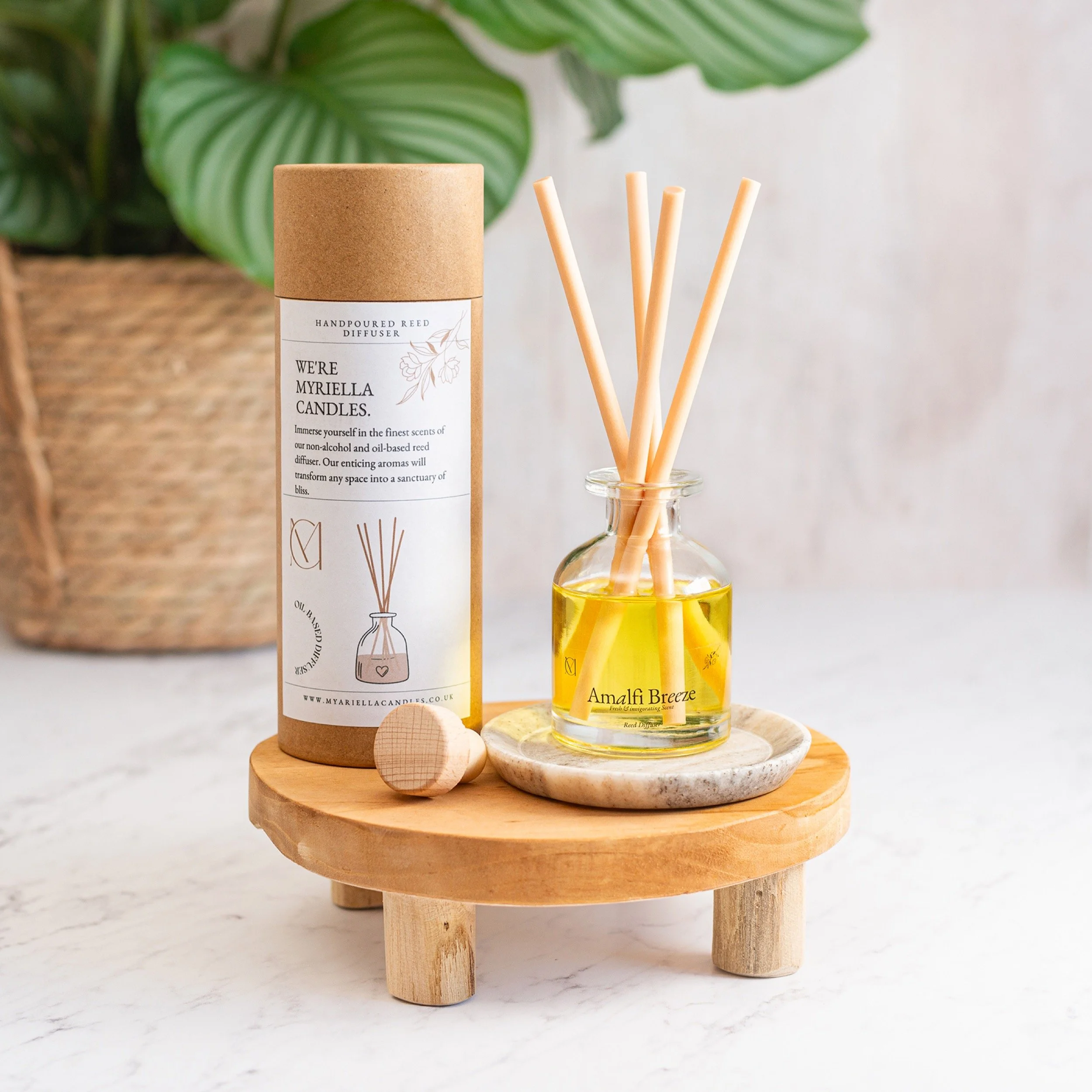 A small wooden tray with diffuser sticks in a glass bottle of yellow oil and a cylindrical brown cardboard box labeled 'We're Myariella Candles,' placed on a white marble surface, with a large green leafy plant in the background.
