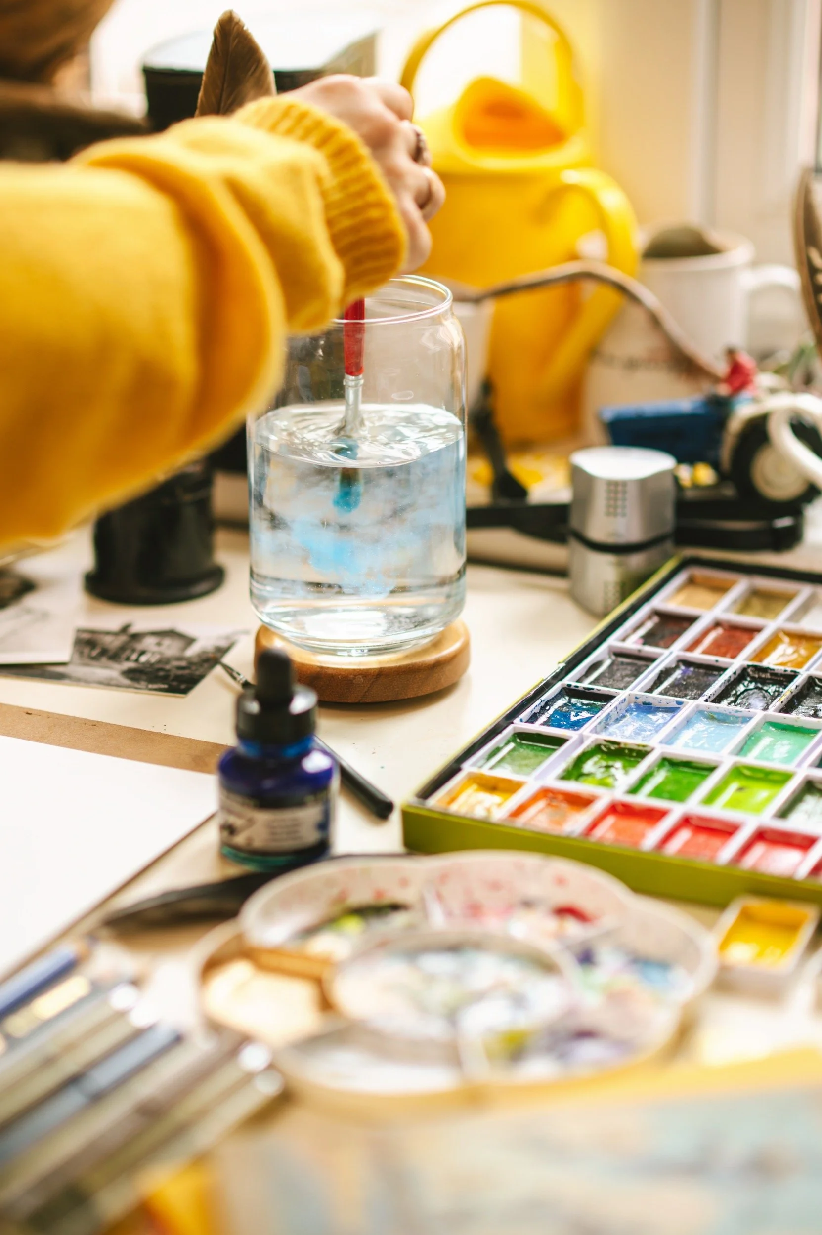 Person stirring water in a glass jar with watercolors, paintbrushes, and art supplies on a cluttered table.