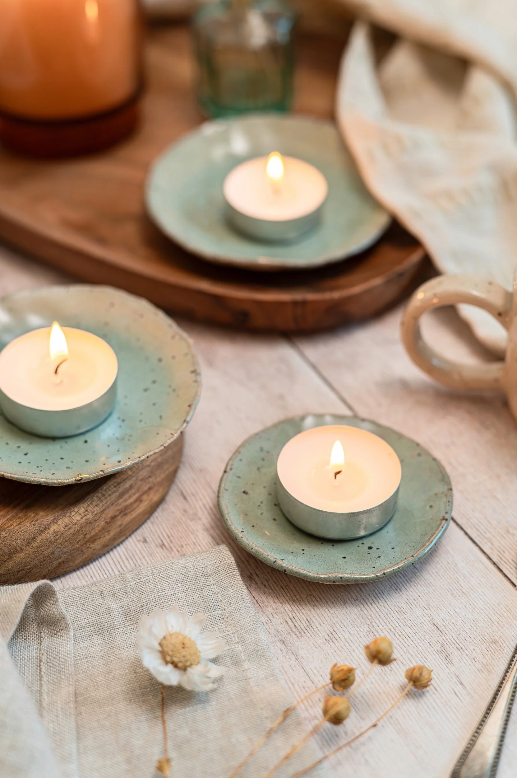 A table setting with three lit tealight candles on green ceramic plates, arranged on wooden trays. There is a beige cloth napkin with a white daisy and dried flowers, and a white ceramic mug in the background.