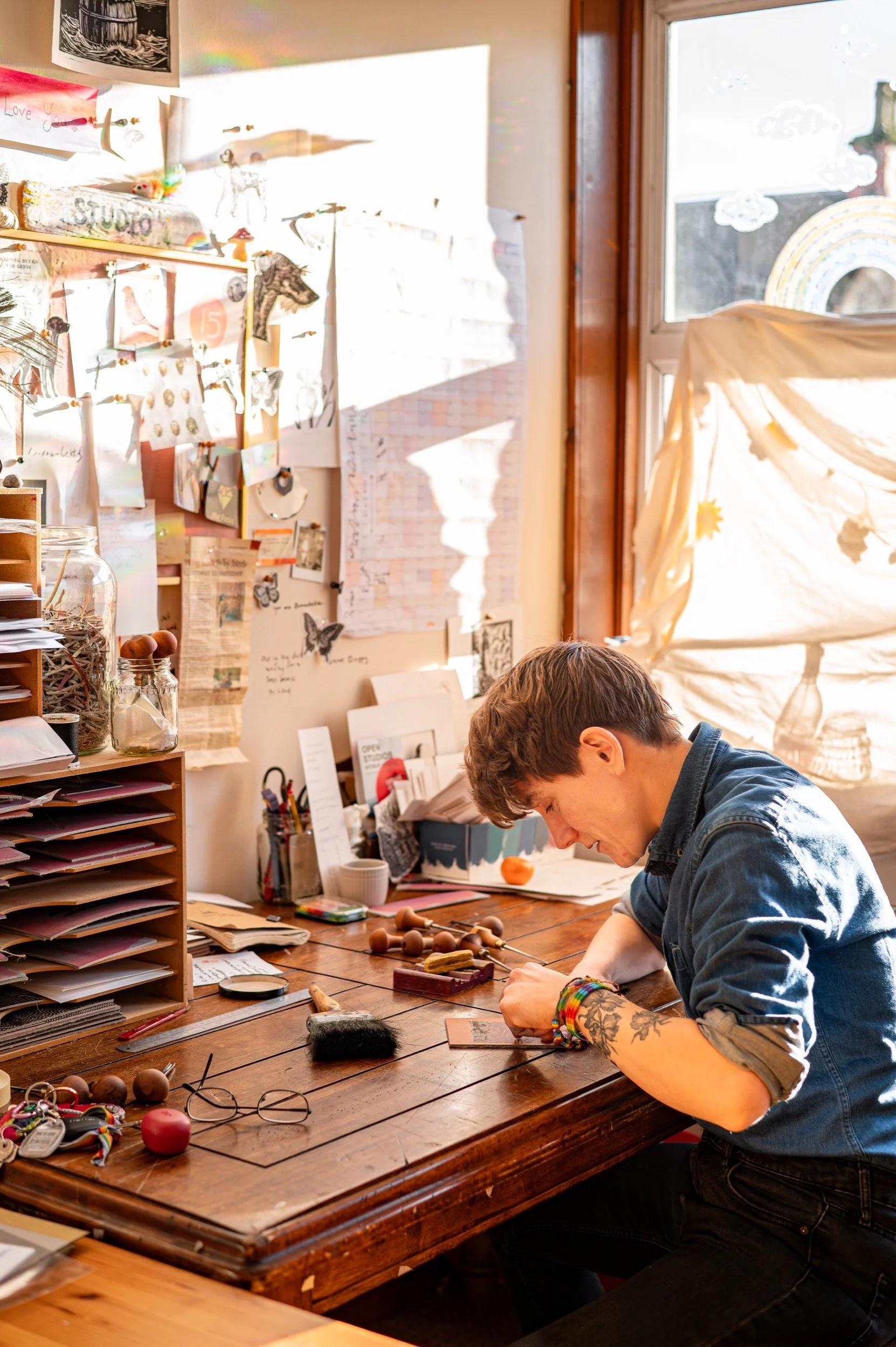 A woman with tattoos wearing a denim shirt, sitting at a cluttered wooden desk, working with small tools. Bright sunlight streams through a nearby window, illuminating a wall filled with papers, drawings, and decorations.