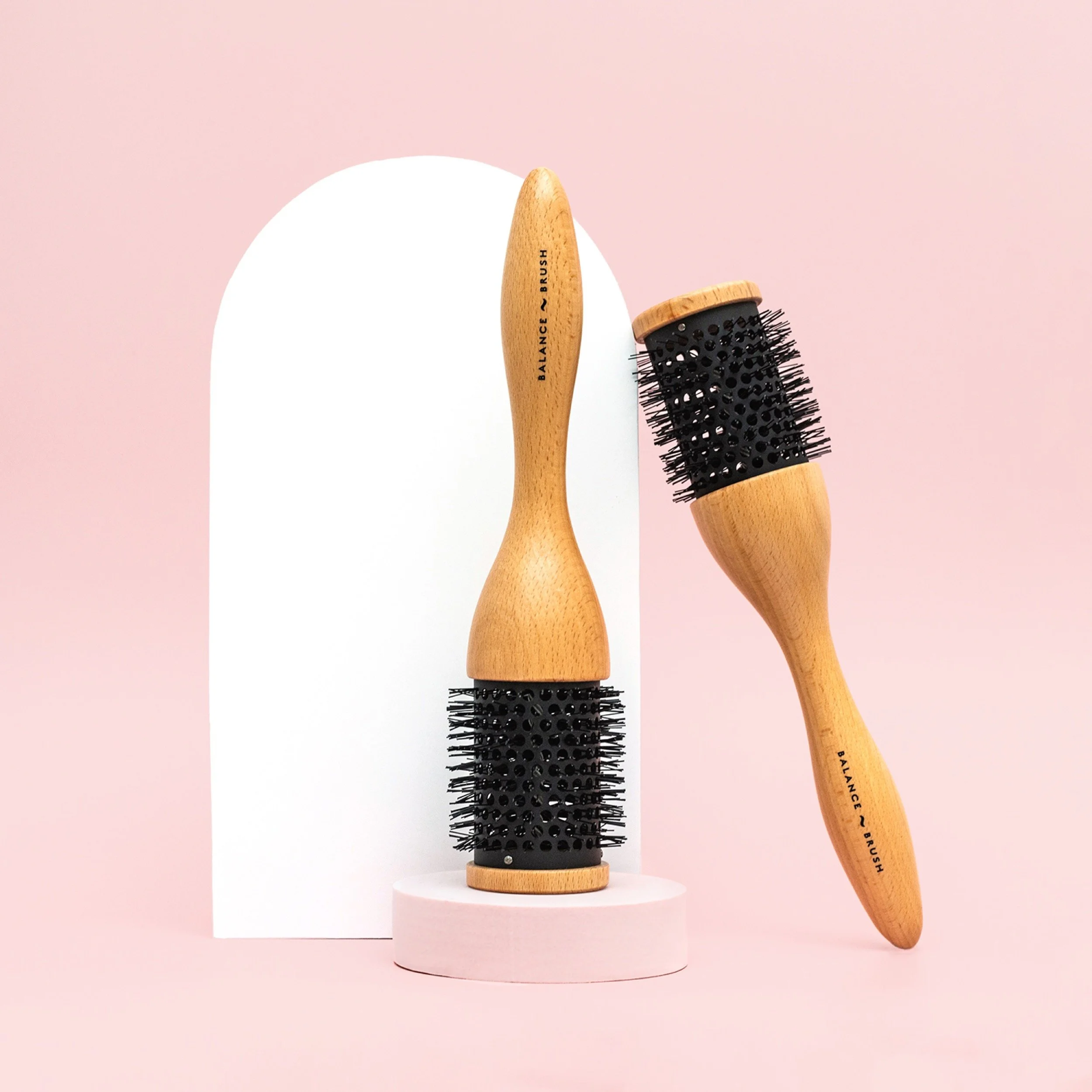 Two wooden handheld brushes with black bristles, one standing upright on a round pink platform and the other leaning against a white arched backdrop, each labeled 'Balance Brush'.